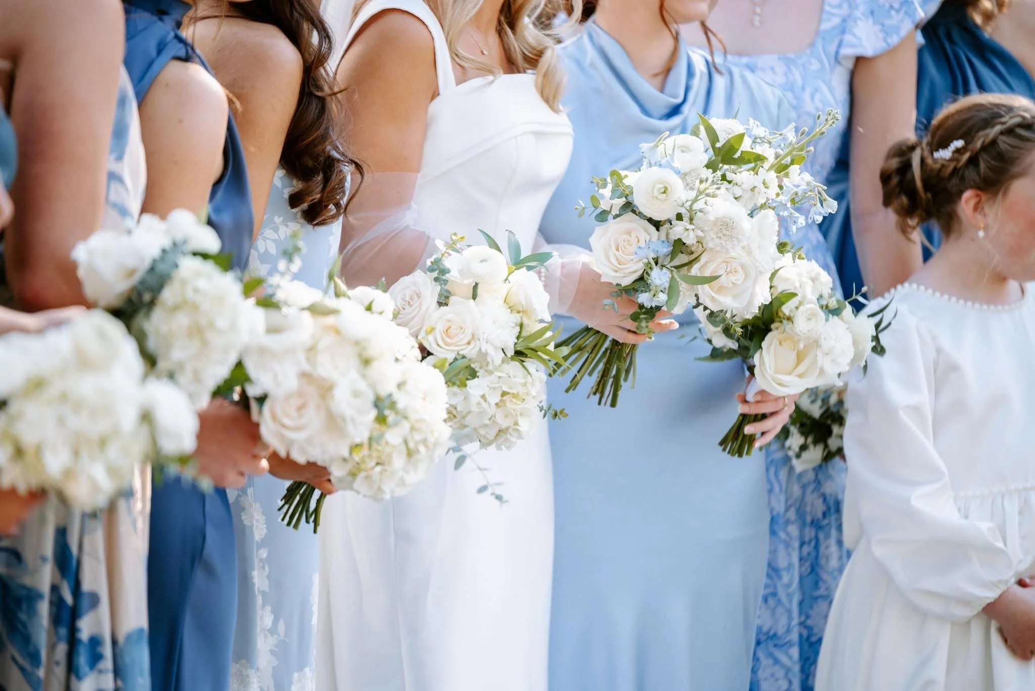 A line of women dressed in pastel blue and white dresses holding bouquets of white flowers during a wedding ceremony.