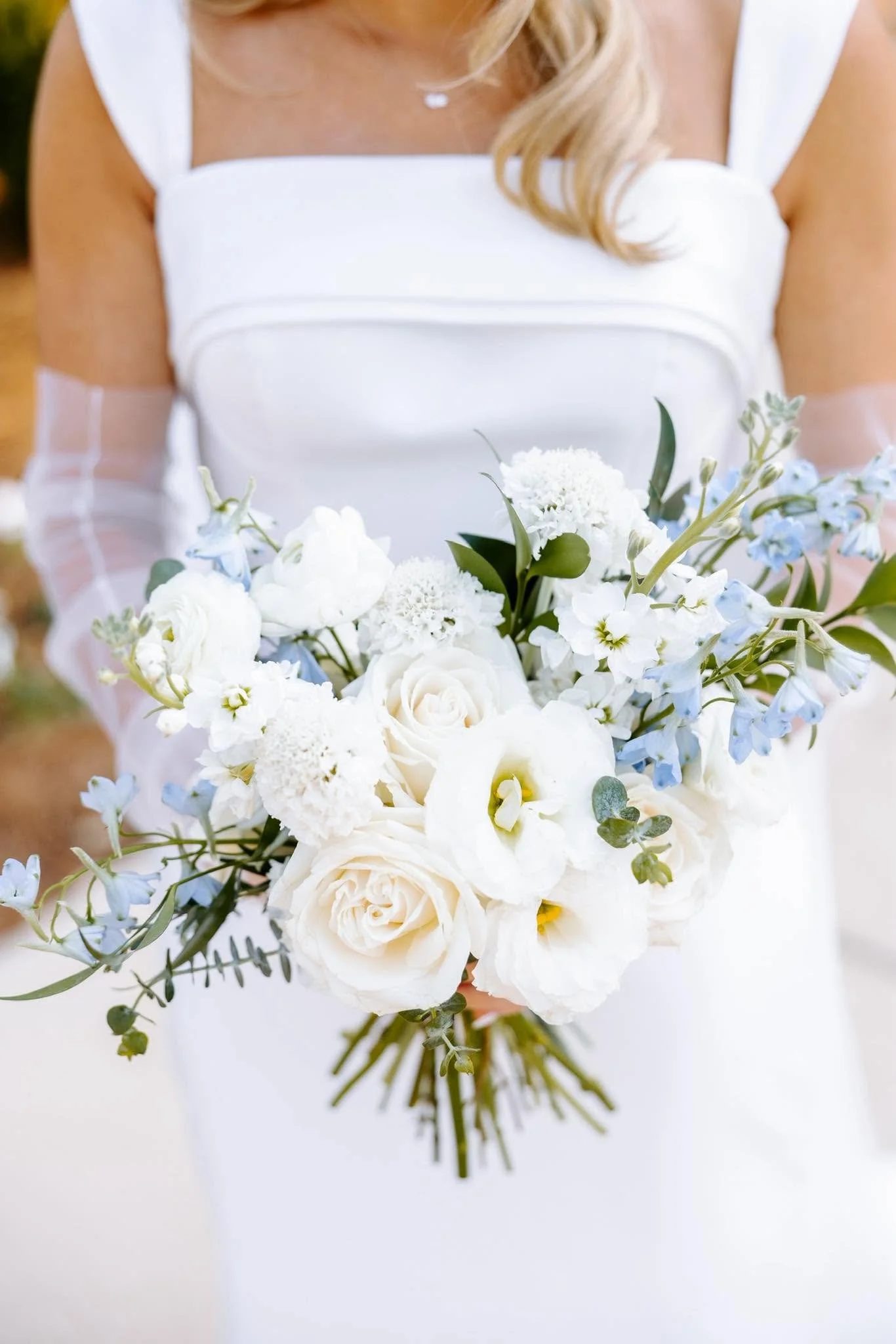 A woman in a white dress holding a bouquet of white and light blue flowers with green leaves.