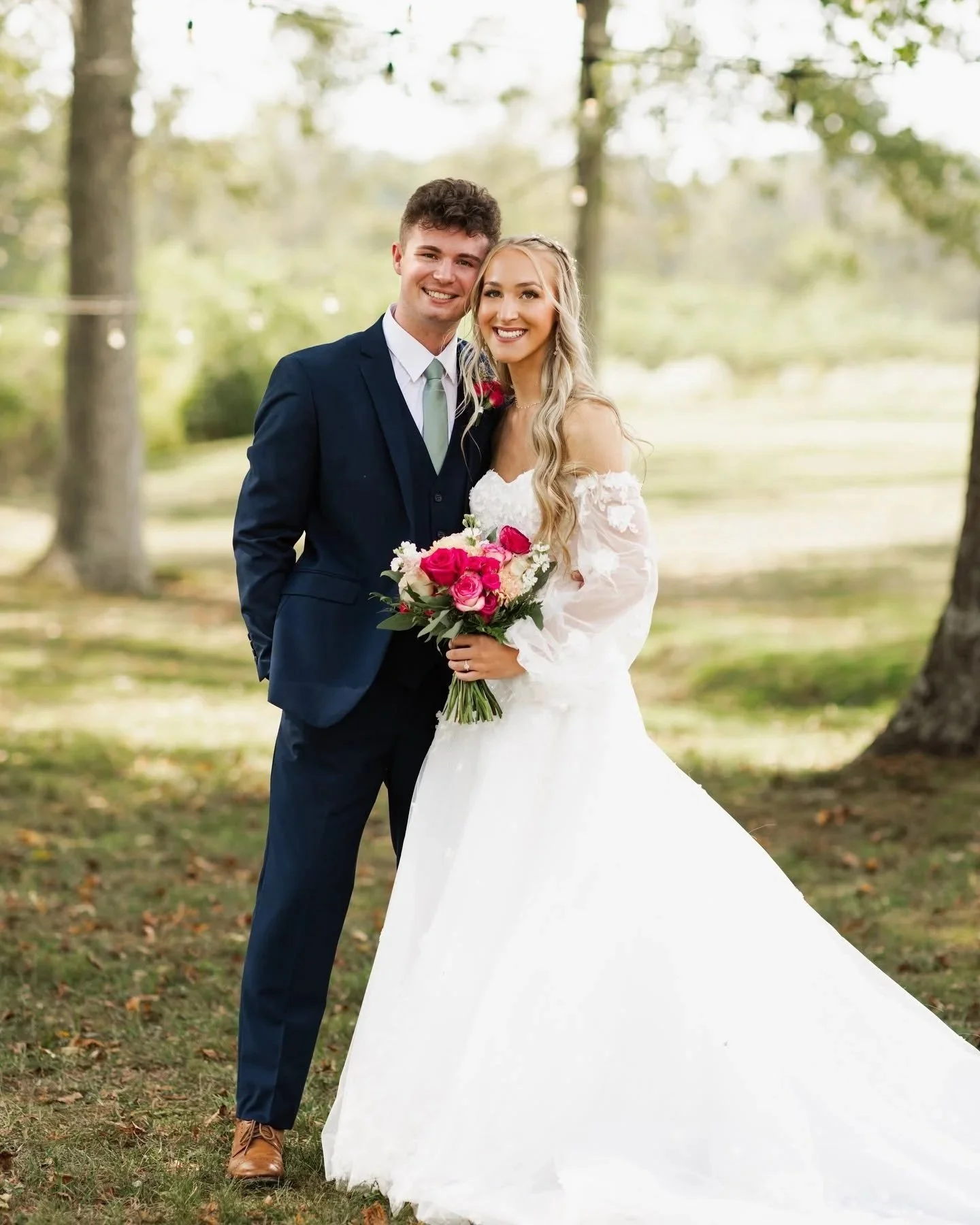 A newlywed couple outdoors in a wooded park, smiling and standing close together. The groom is dressed in a navy suit, white shirt, and light green tie, while the bride wears a white wedding dress with lace details and holds a bouquet of pink and red roses. They are surrounded by trees and fall foliage.