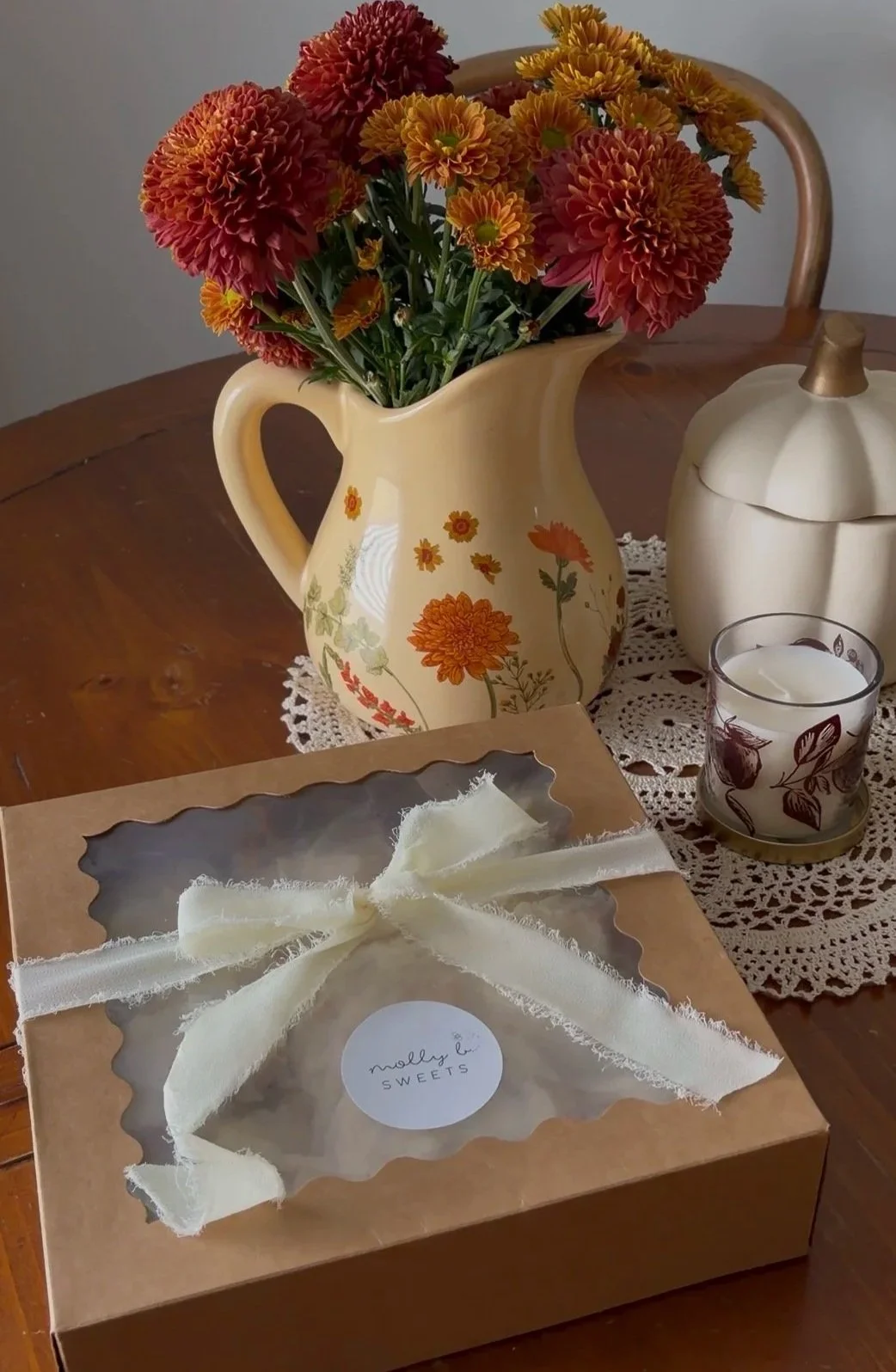 A flower arrangement of orange and yellow flowers in a cream-colored pitcher with floral patterns sits on a dining table. Beside it is a cream-colored pumpkin-shaped container, a lit candle in a glass holder with floral design, and a gift box with a white ribbon on a lace doily.