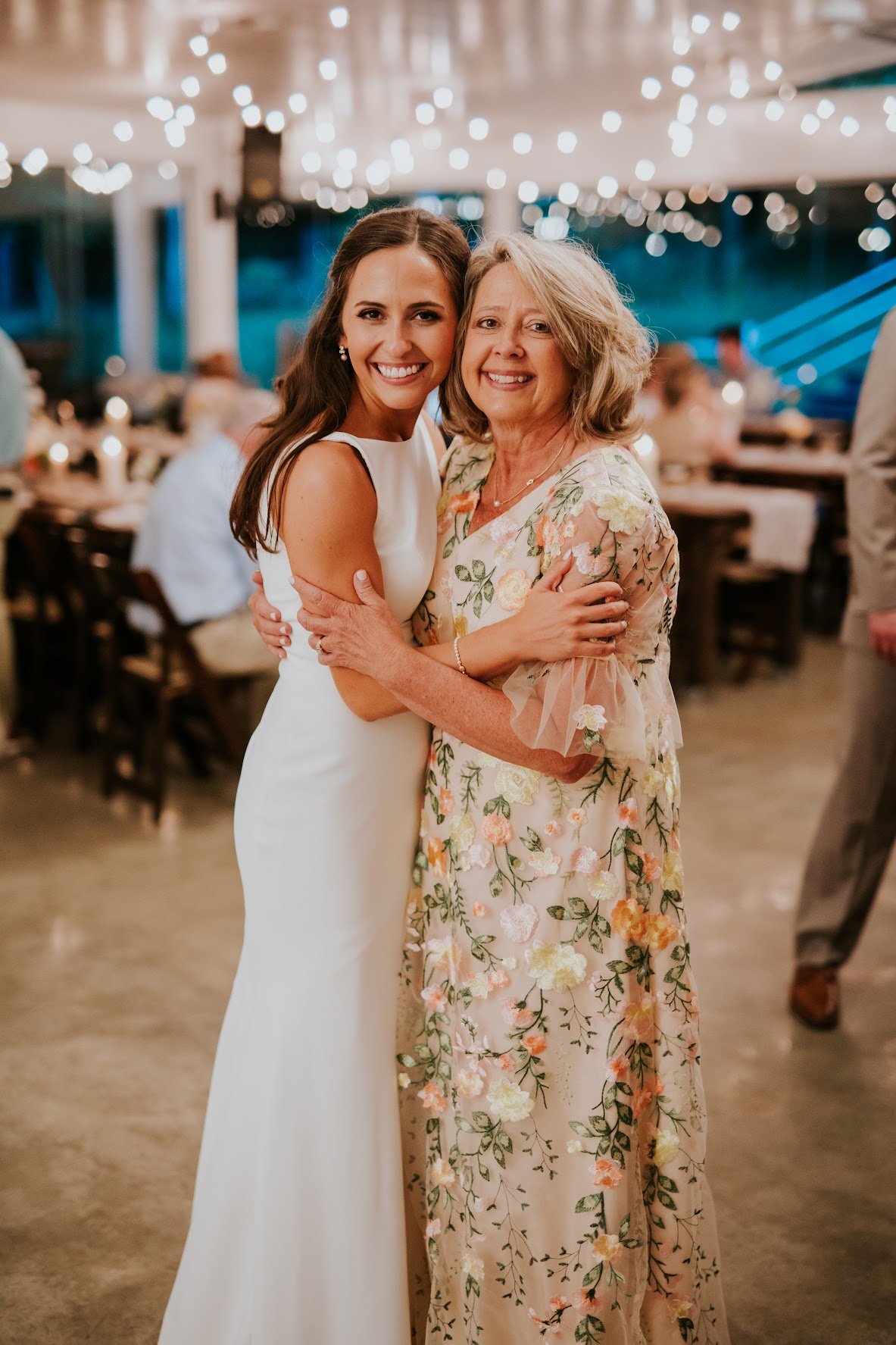 A woman in a white wedding dress hugging an older woman in a floral dress at a wedding reception.