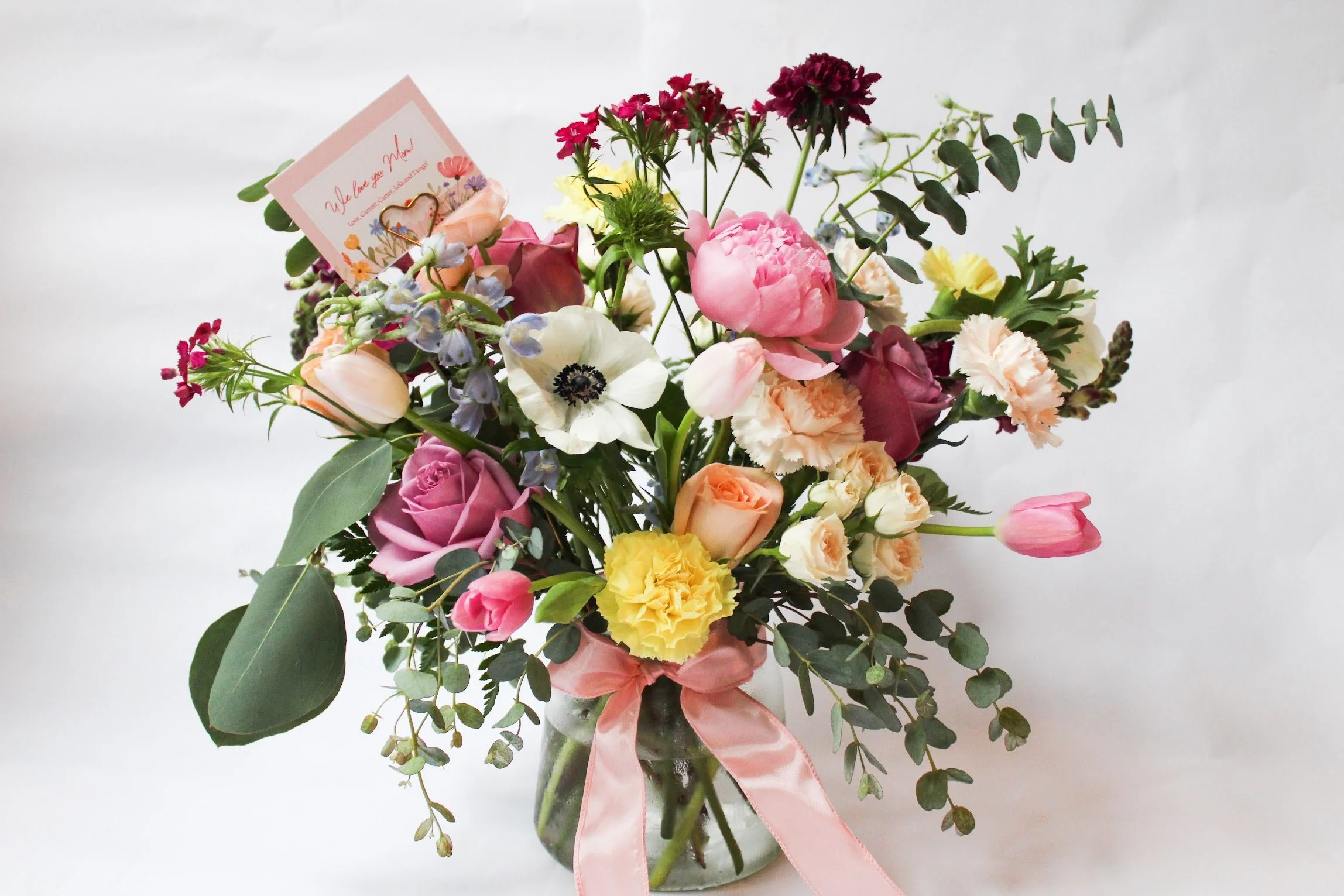 A bouquet of mixed flowers including pink roses, yellow carnations, tulips, anemones, and greenery, tied with a pink ribbon, with a greeting card attached.