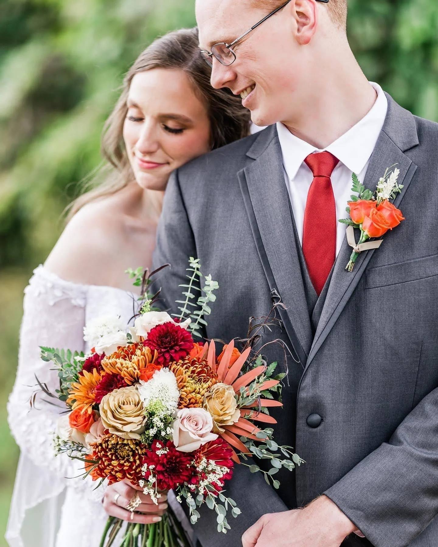 A bride and groom at their wedding, with the bride holding a colorful bouquet of flowers and the groom wearing a gray suit with a red tie and boutonniere, outdoors with blurred greenery in the background.