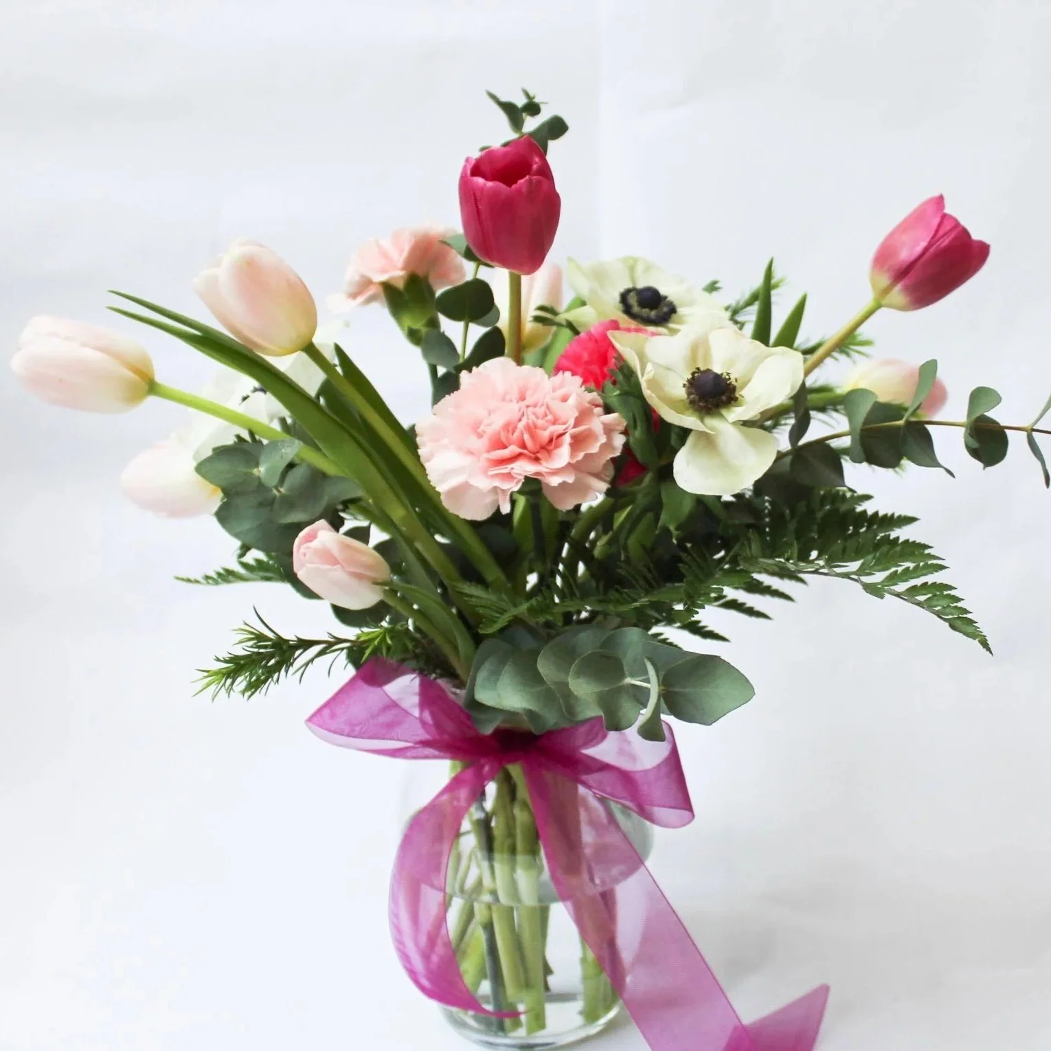 A bouquet of pink, white, and red flowers tied with pink ribbon in a glass vase.