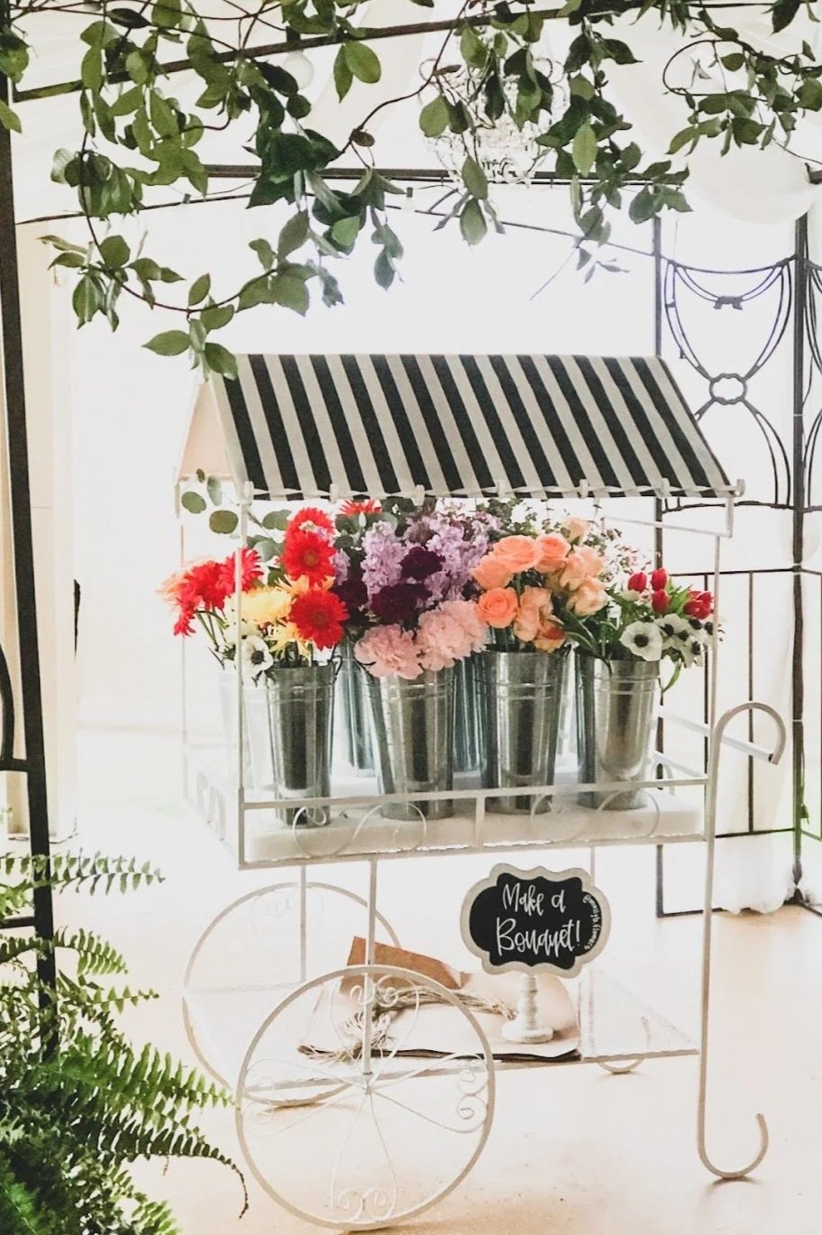 A decorative flower cart filled with colorful flowers in metal buckets, with a sign that says 'Make a Bouquet' at the front, and greenery hanging overhead.