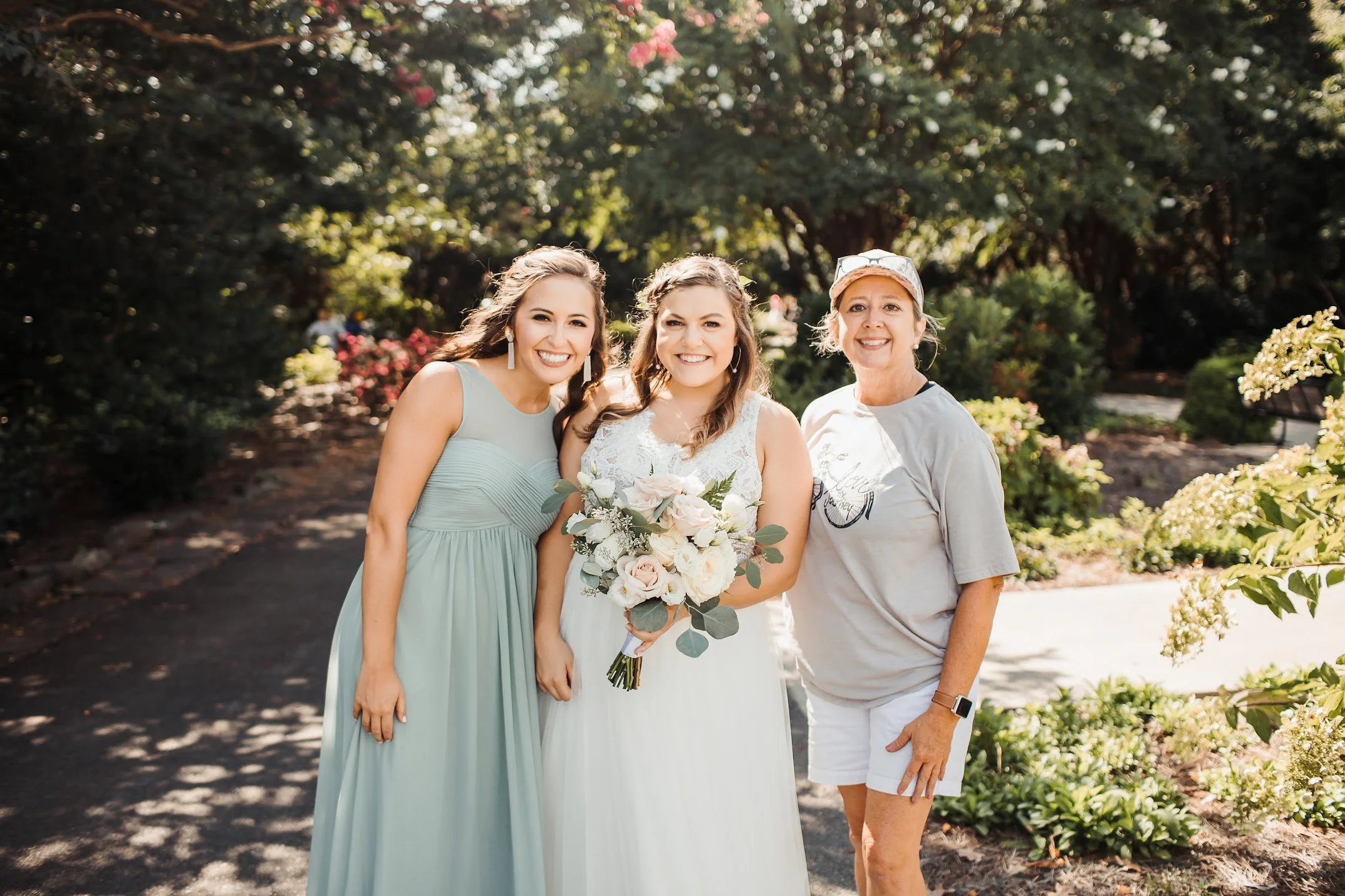 Three women smiling at the camera outdoors during daytime, one holding a bouquet of white and pink flowers, standing among trees and bushes.