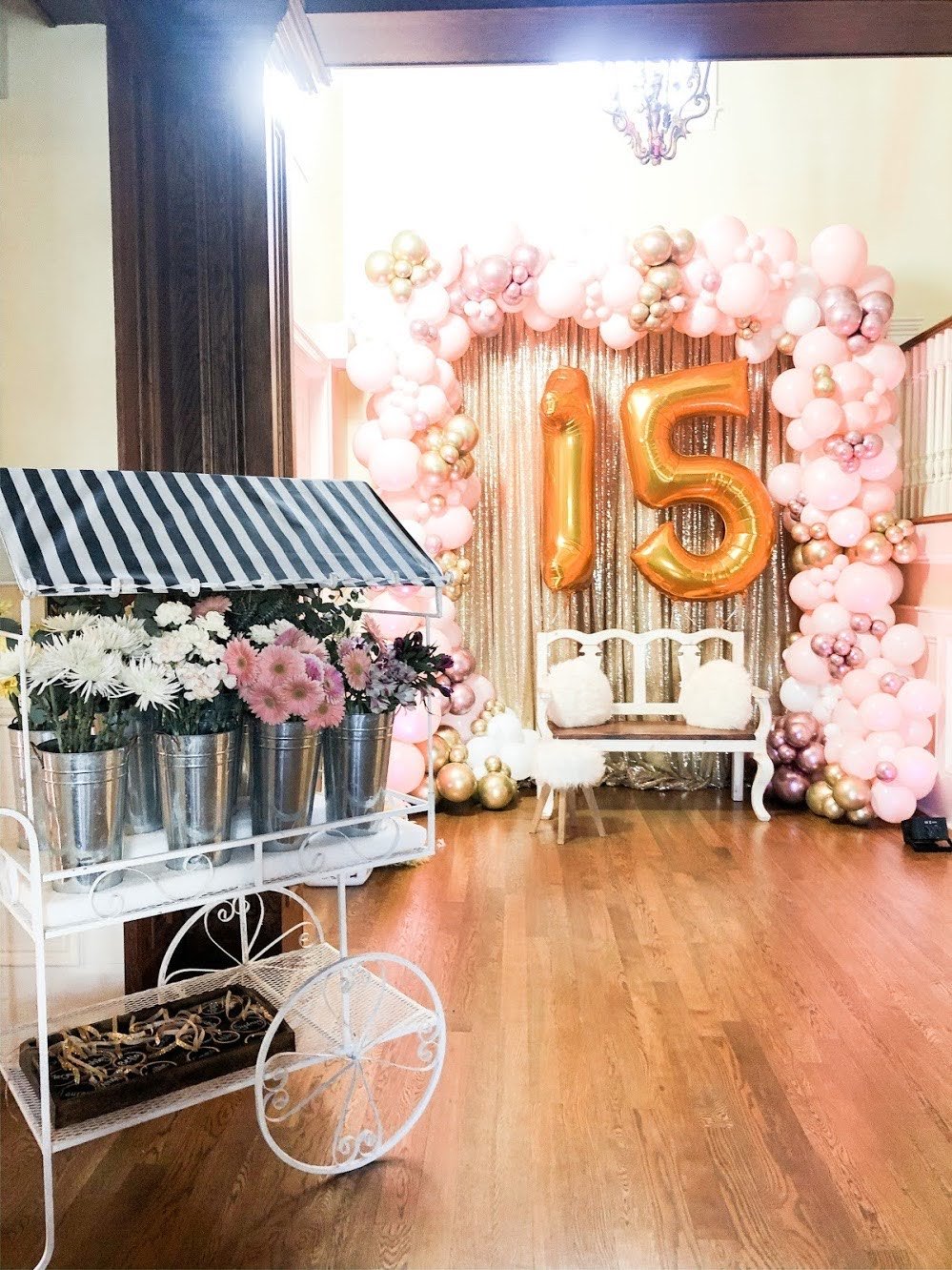 Decorative setup with gold number balloons '15' surrounded by pink, white, and gold balloons, a vintage wooden bench, and a balloon arch, likely for a 15th birthday celebration.