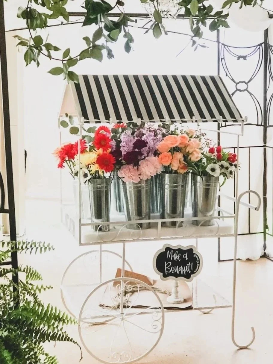 A decorative garden cart displaying colorful flowers in metal buckets with a black and white striped canopy and a sign that reads 'Make a Bouquet!'