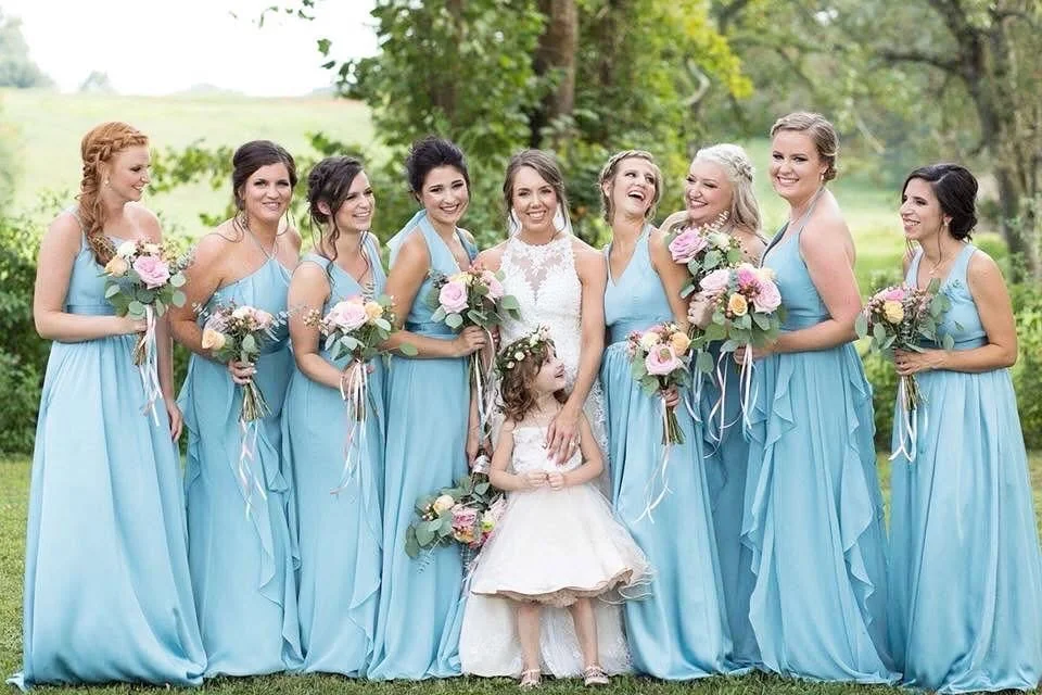 Bride and her bridesmaids in blue dresses standing outdoors, holding bouquets, with a young girl in a white dress in front.