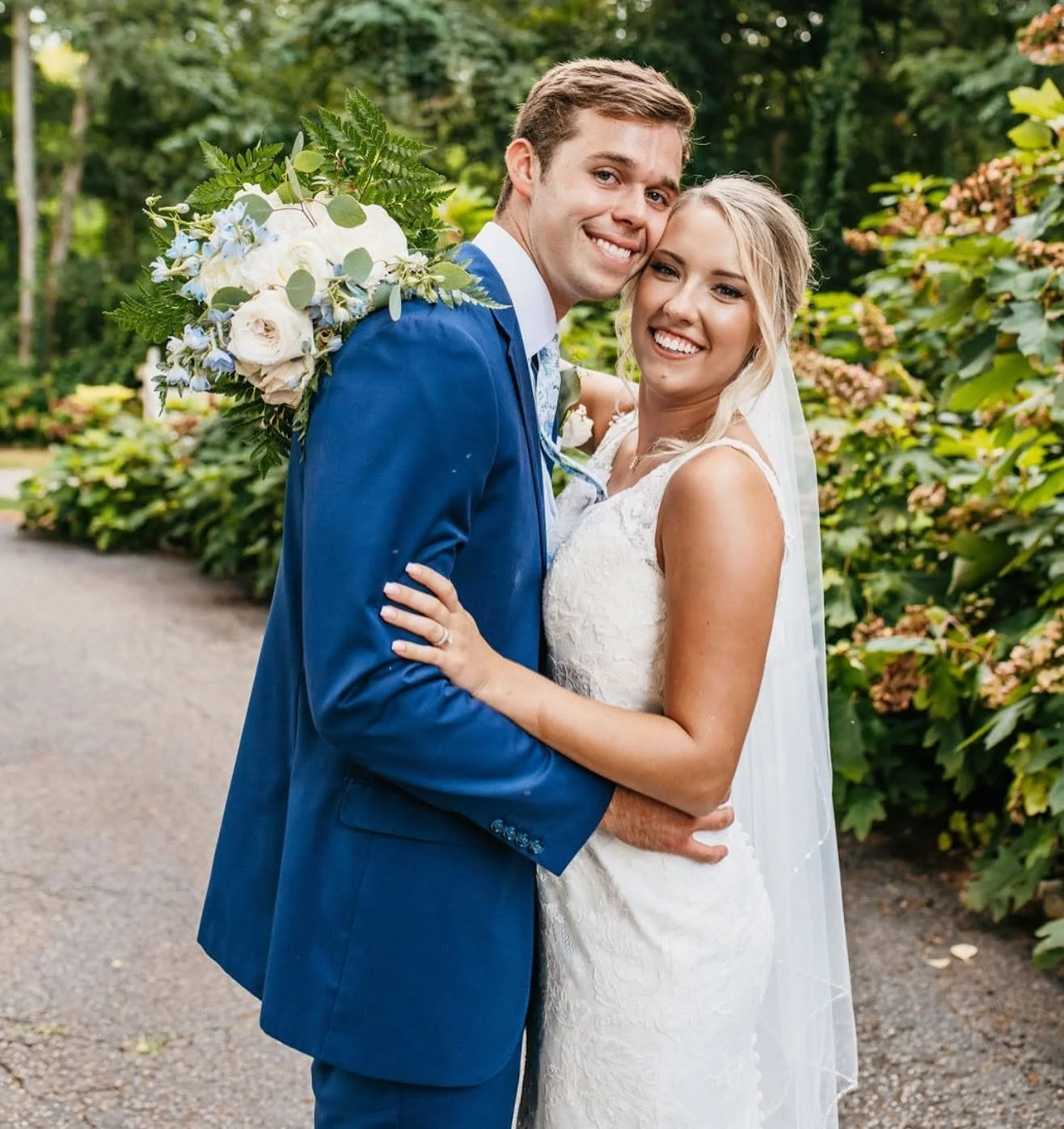 A joyful newlywed couple smiling and embracing outdoors, with greenery and flowering bushes in the background.