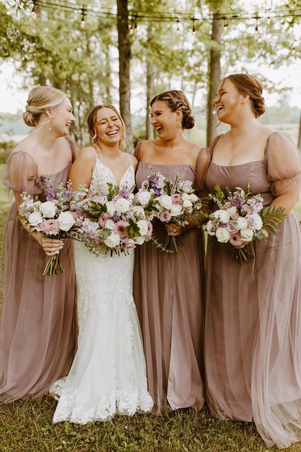 A bride and three bridesmaids standing together outdoors, all holding bouquets of pink and white flowers, laughing and smiling among trees.