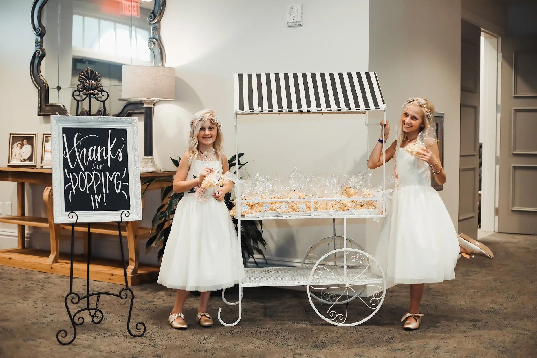 Two young girls dressed in white dresses with floral headbands standing beside a vintage-style white cotton candy cart with black and white striped canopy. One girl is holding a bag of cotton candy, and the other is holding a stick of cotton candy. T