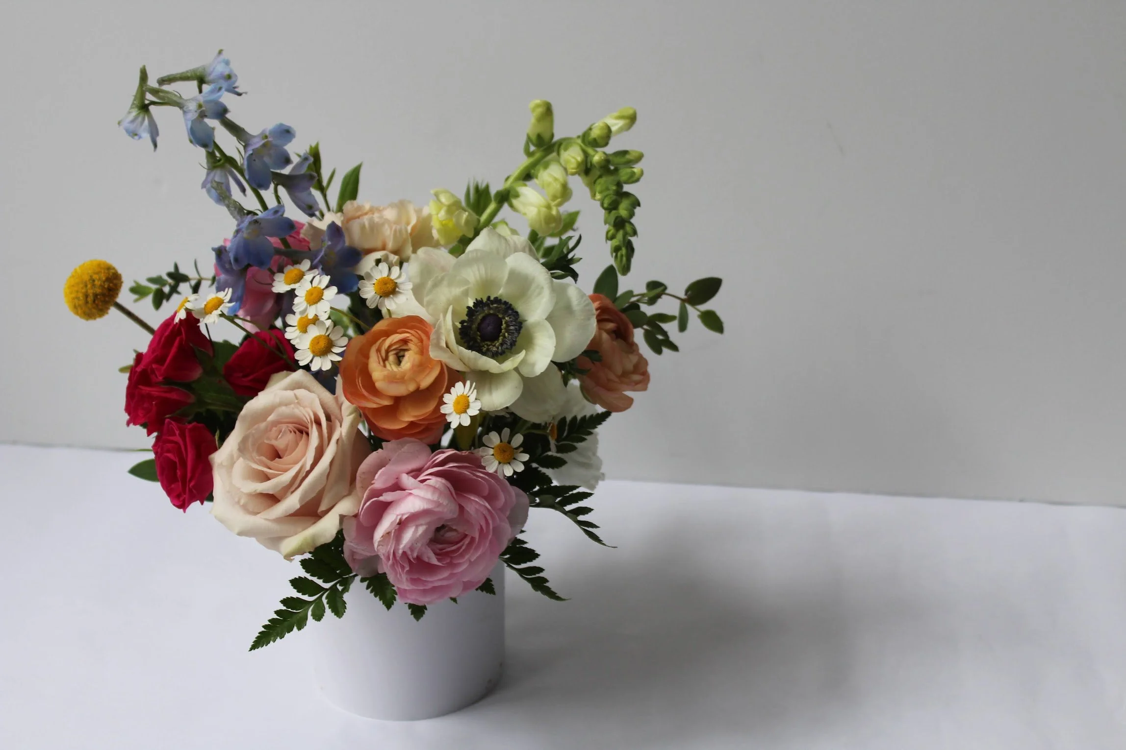 A bouquet of multicolored flowers in a white vase against a plain light-colored background.