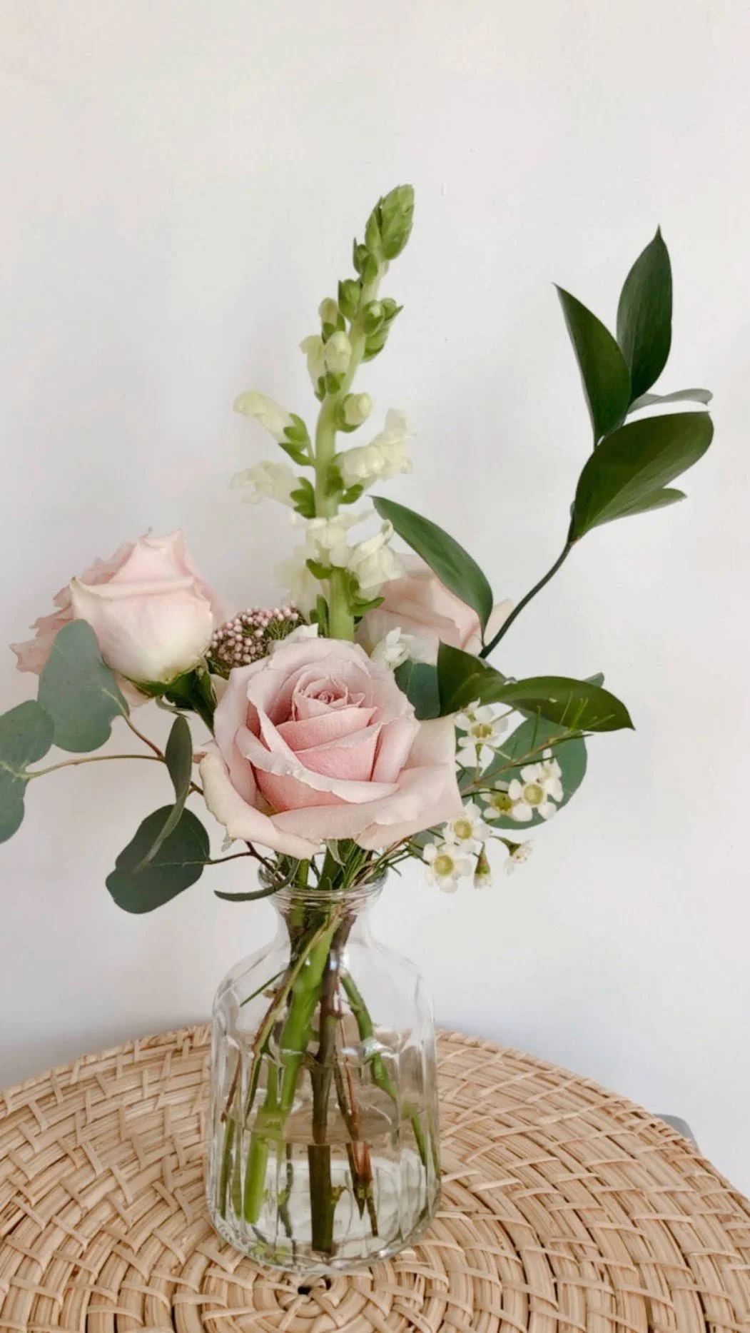 A small bouquet of flowers in a clear glass vase on a woven rattan table. The bouquet includes pale pink roses, white stock flowers, small white filler flowers, and green leaves, all set against a plain white background.