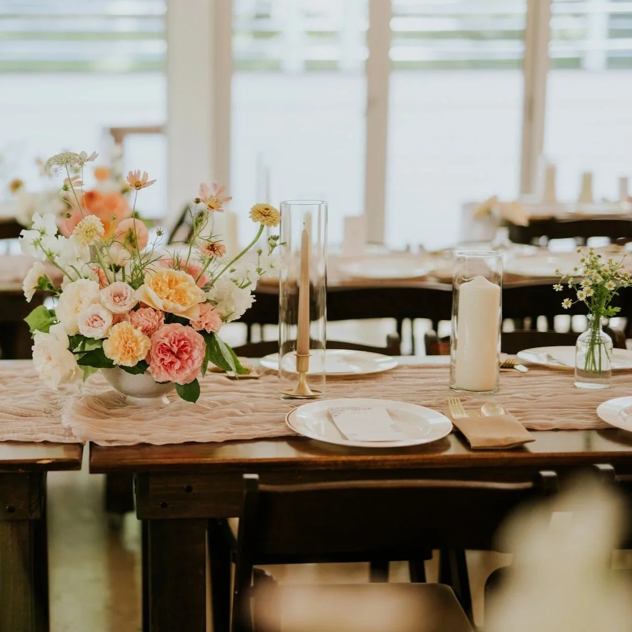 Elegant dining table decorated with a bouquet of pink, cream, and peach flowers, a tall glass candle holder with a candle, a smaller vase with daisies, and a white candle in a glass holder, set for a special occasion in a bright room.