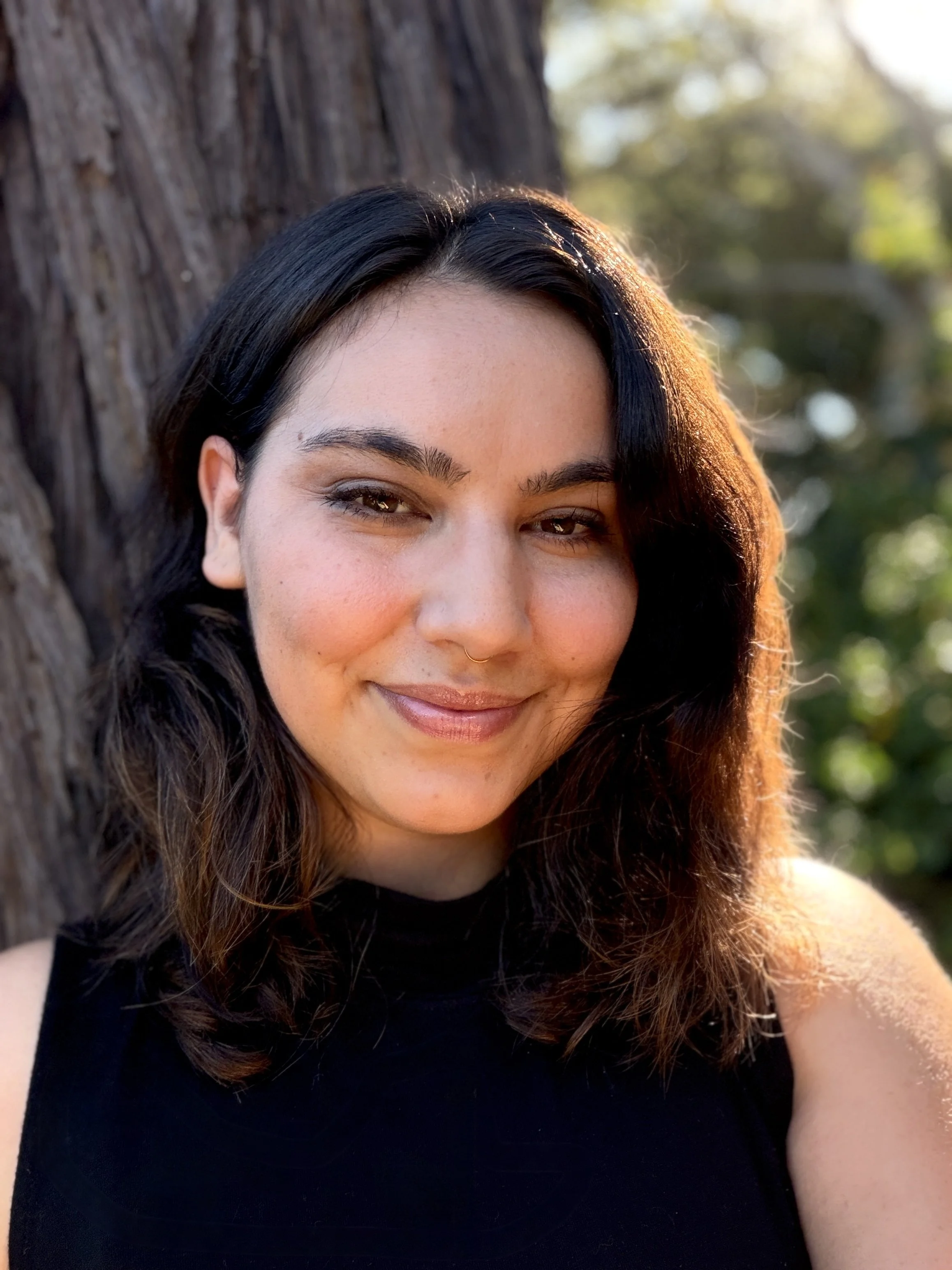 Close-up of a woman with dark, wavy hair smiling outdoors by a tree in sunlight.