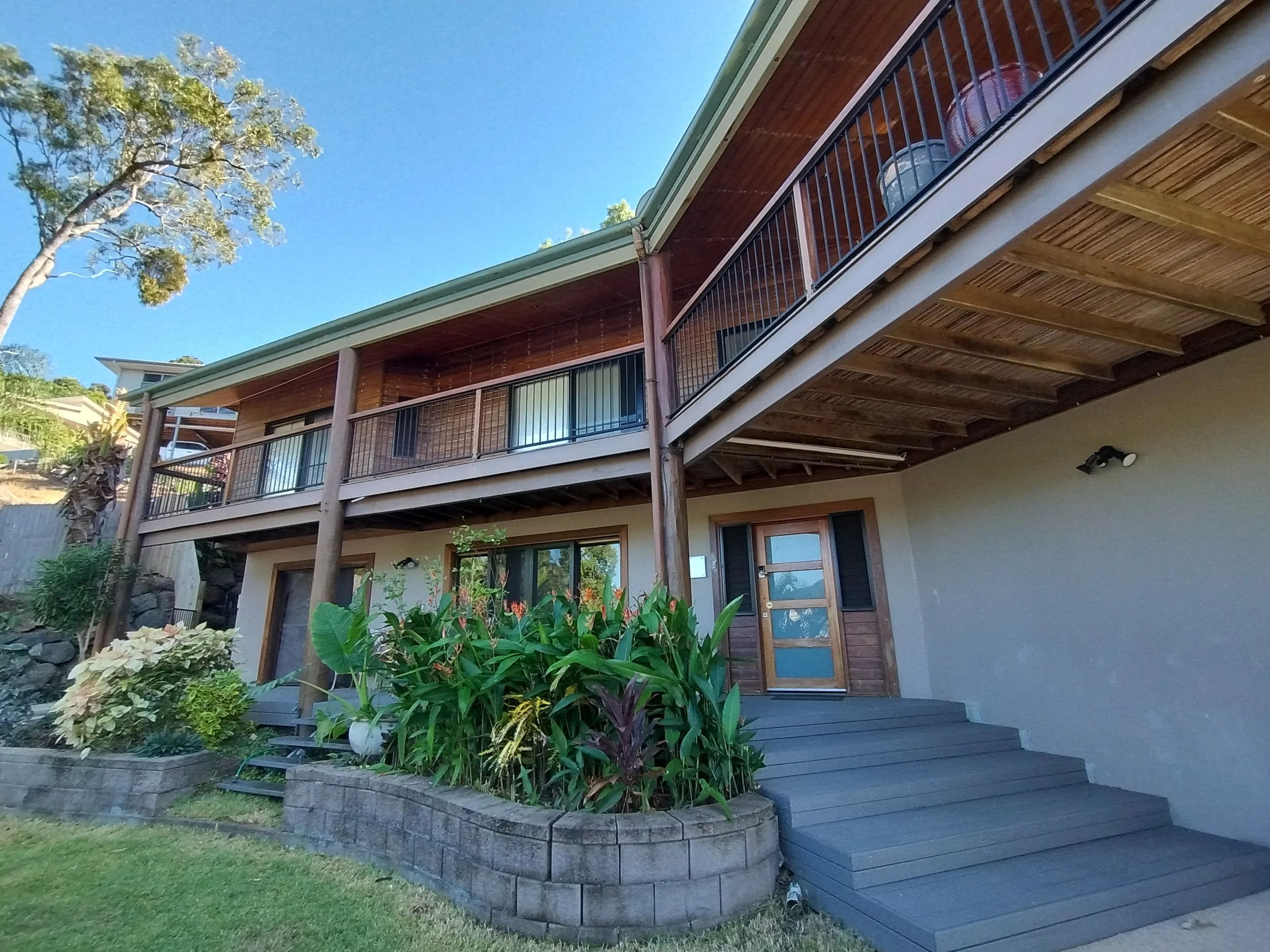 Backyard view of a two-story house with wooden siding, a balcony with a metal railing, and landscaped garden with various plants and flowers under a clear blue sky.