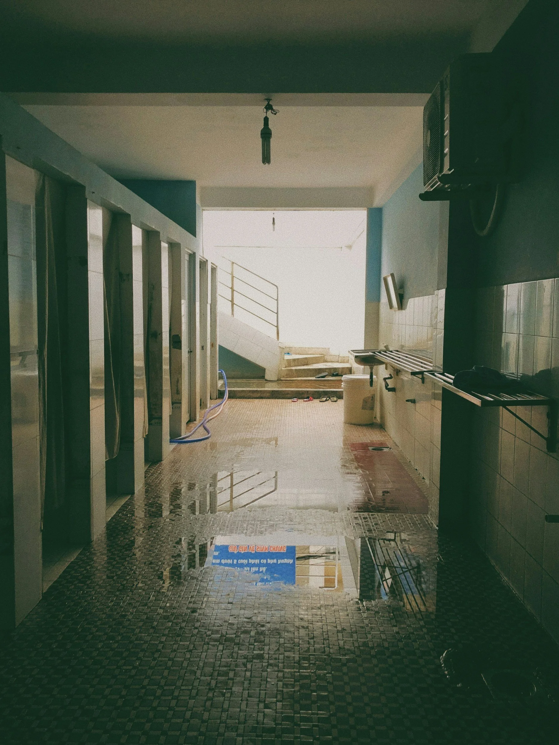 An indoor corridor with a tiled floor, partially flooded with water. The corridor leads to an open staircase. There are some items on the floor, a blue hose, a white bucket, and a sign reflected in the water. The ceiling has a hanging light fixture. To the right, there is a black gas heater and a metal rack.