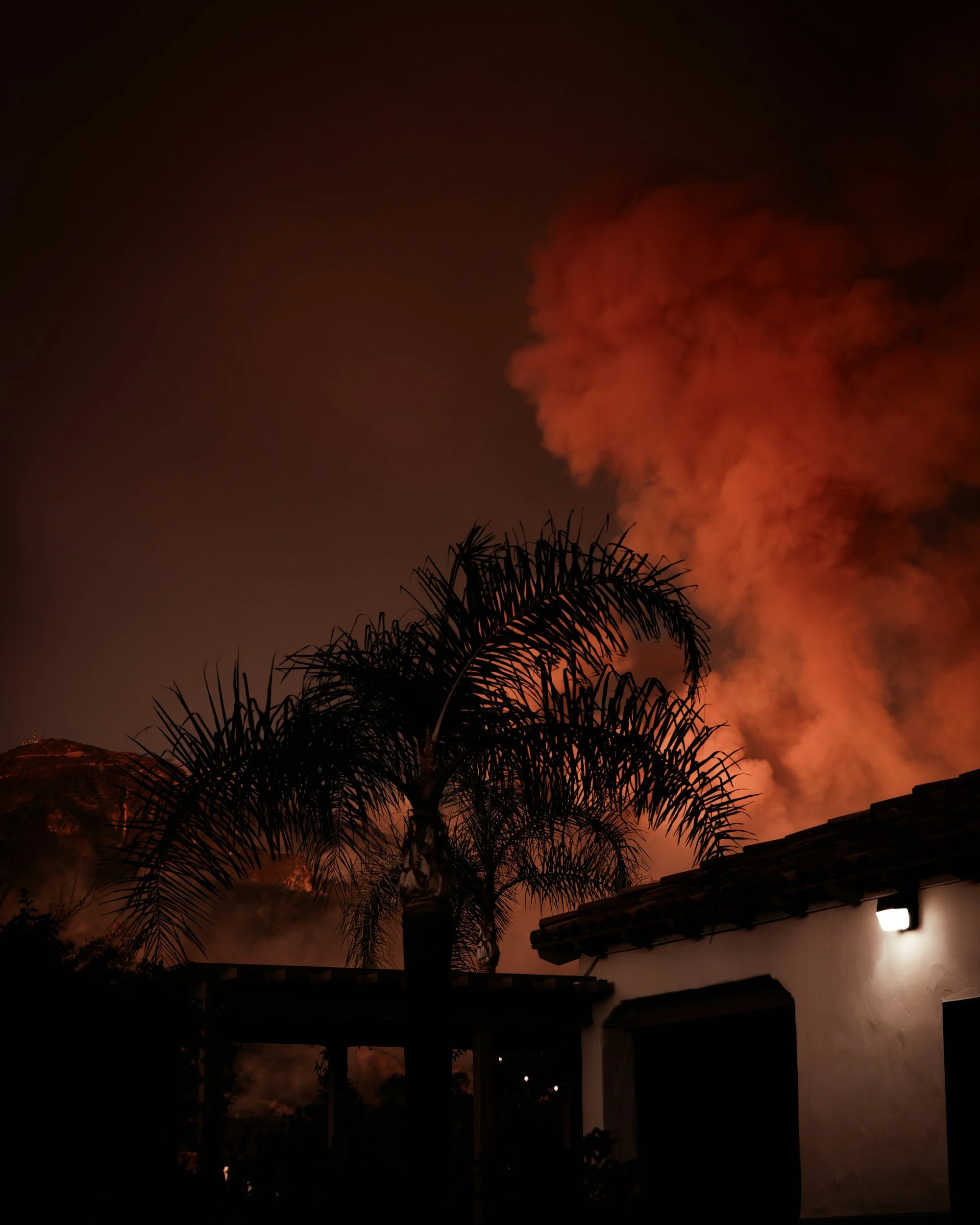 Nighttime scene with orange-tinted clouds in the sky, silhouette of palm trees, house roof, and a small illuminated wall light.