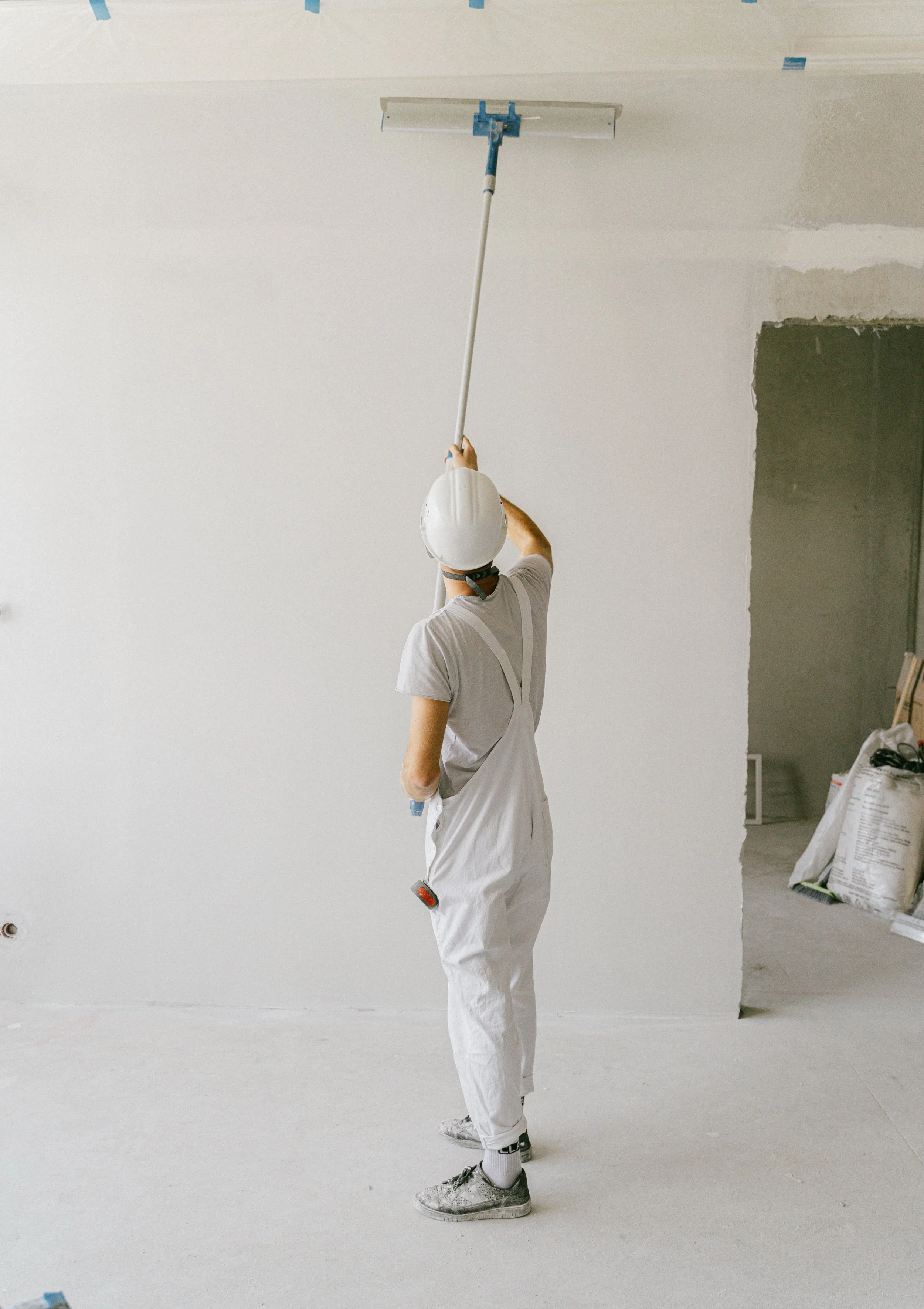 Person painting a wall using a roller extension pole, wearing a white helmet, gray t-shirt, white overalls, and sneakers, in an unfinished room with construction supplies.