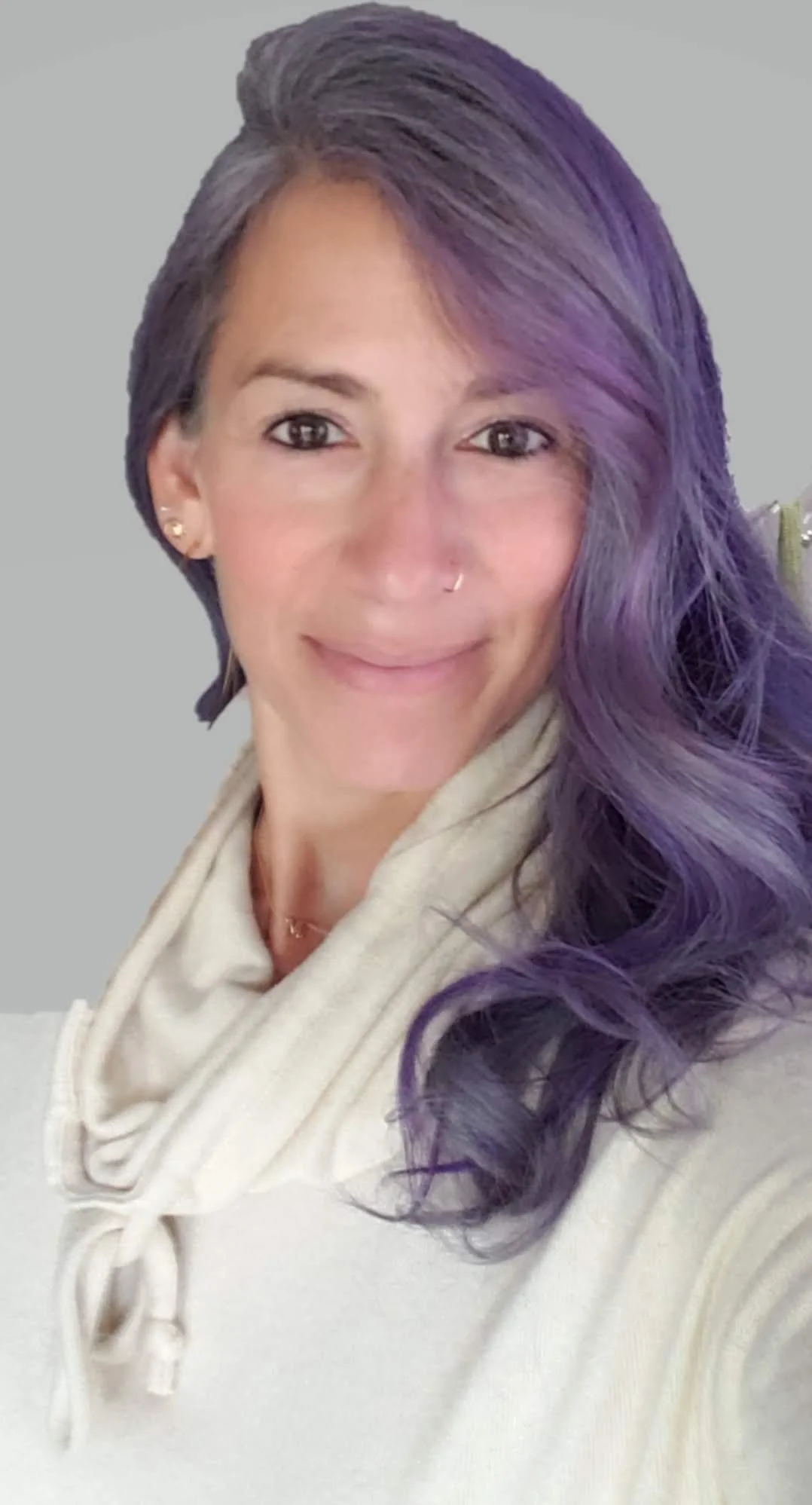 A woman with gray and purple hair styled in waves, smiling with light makeup, wearing gold earrings and a nose ring, dressed in a beige scarf and cream-colored sweater against a plain background.
