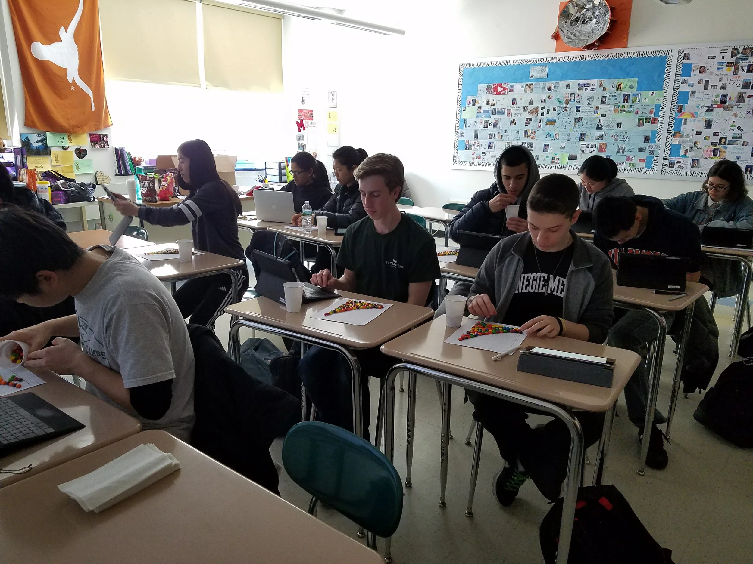 Students in a classroom working on a project with Skittles candies on paper, some students are using cell phones, and a teacher's desk is visible with supplies and a large flag on the wall.