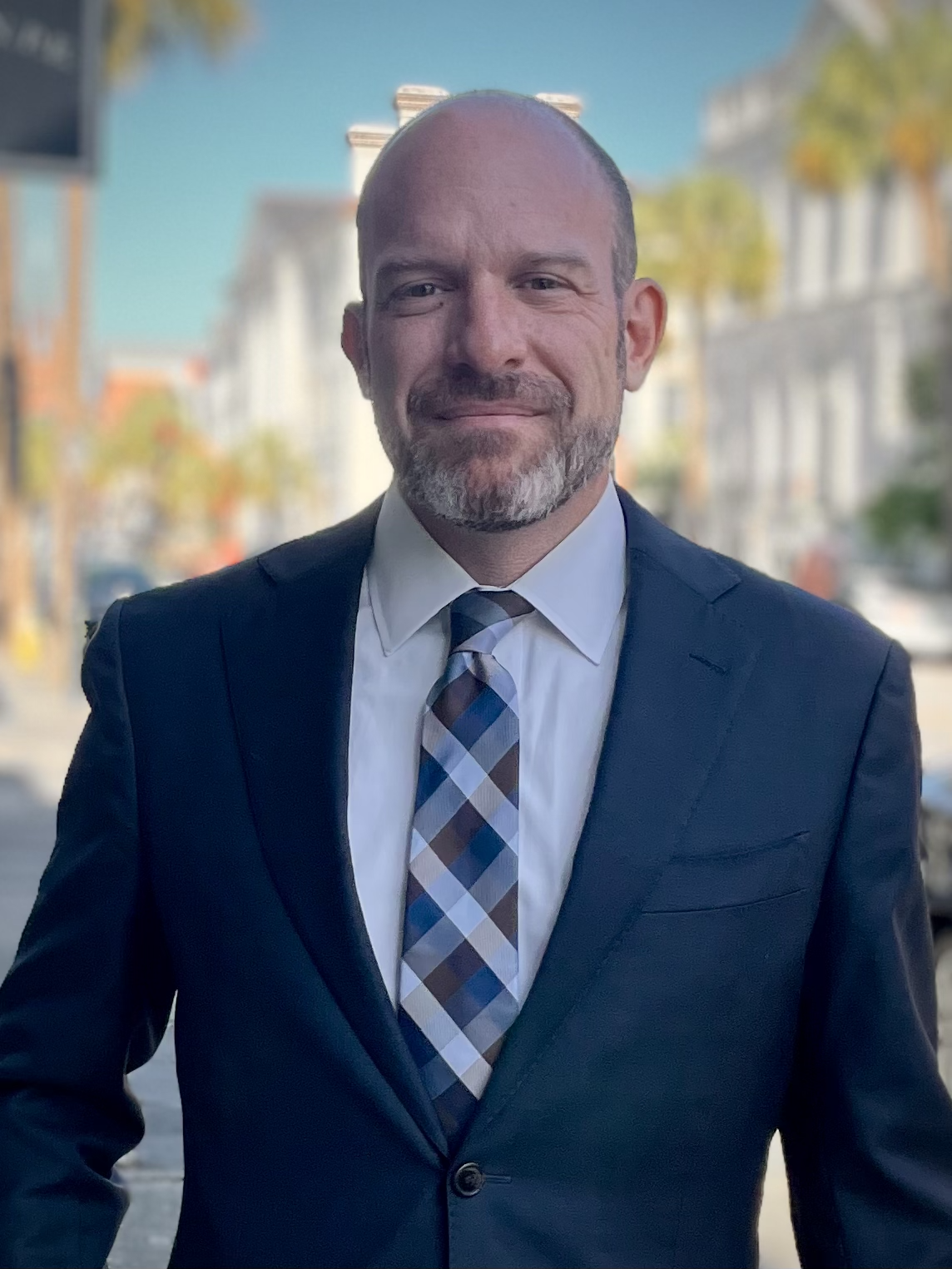 A man in a suit and tie standing outdoors with a blurred city street background.