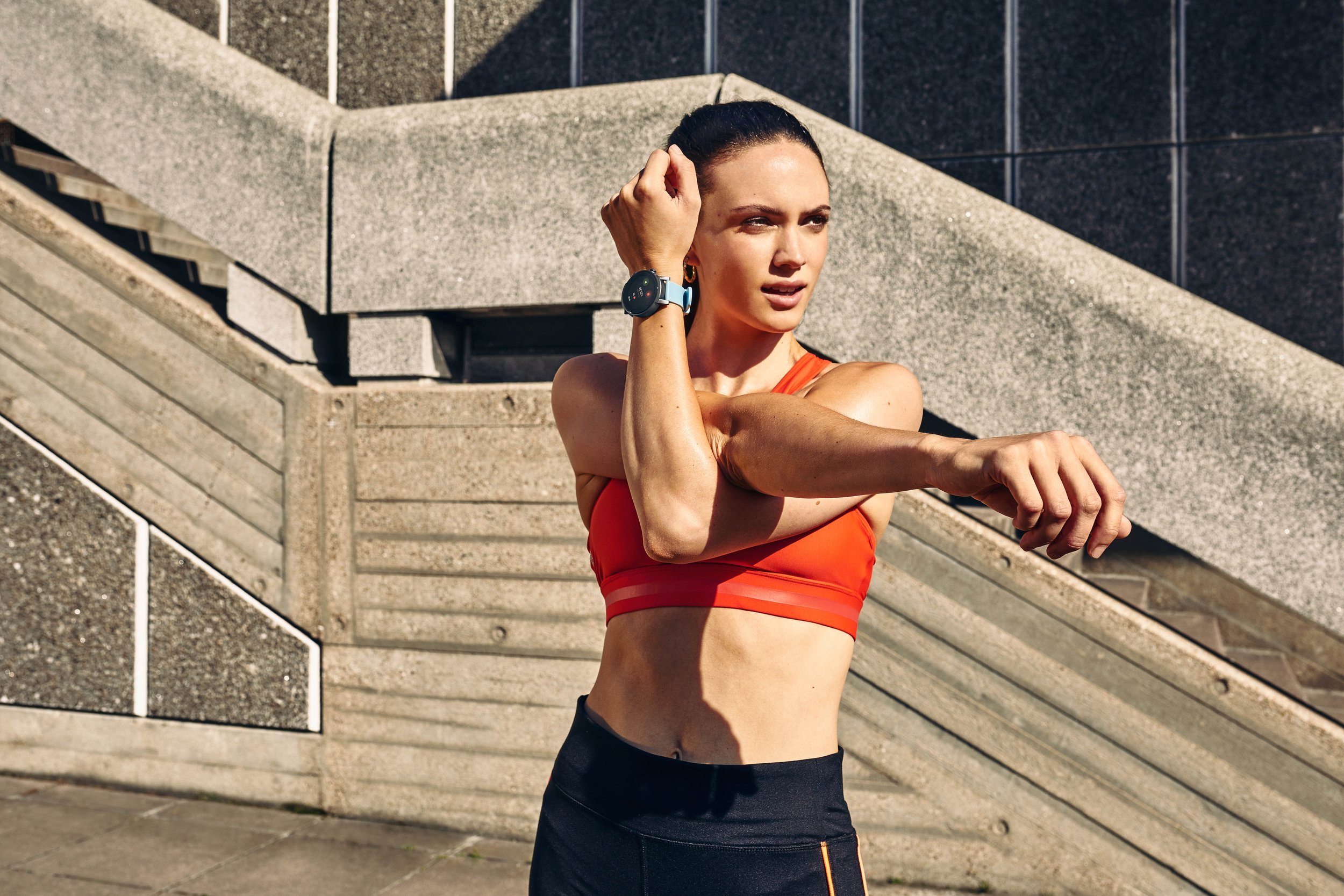 A young women wearing a smart watch warms up before a run in the heart of the city. She wears sports clothing in this Lifestyle photography by Jon Enoch.