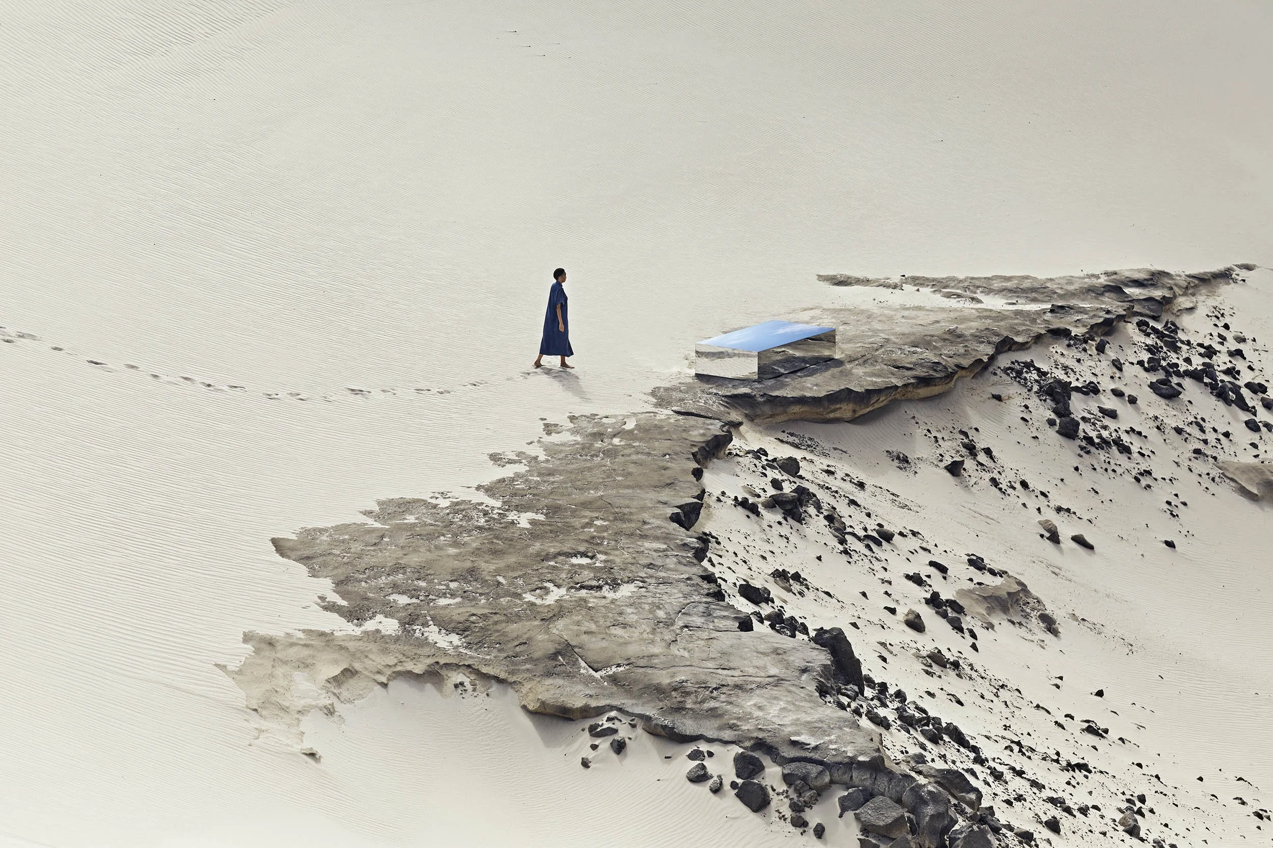 A lone figure walks across the sand dunes towards a mirrored structure in the Cape Town landscape.