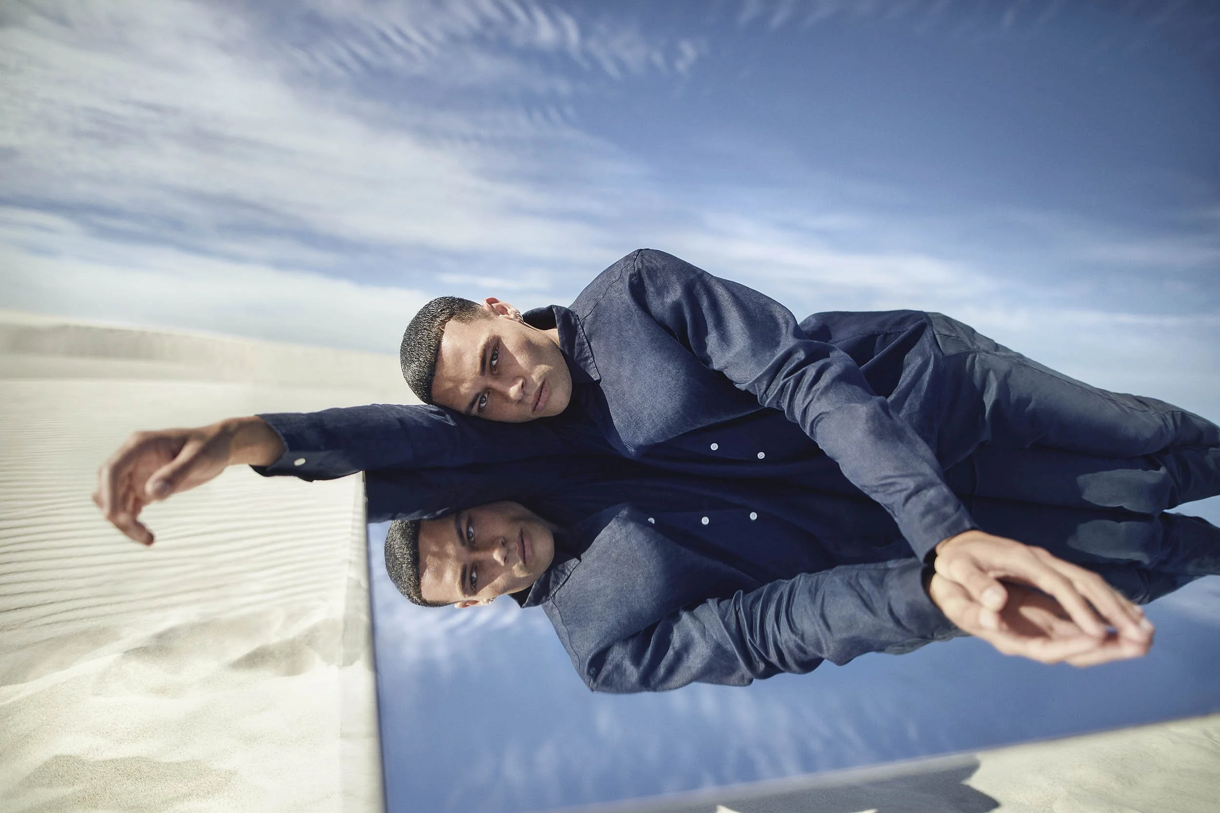 Model reclining on a mirrored structure in the Cape Town dunes, creating a reflection that blends with the surrounding landscape and sky.
