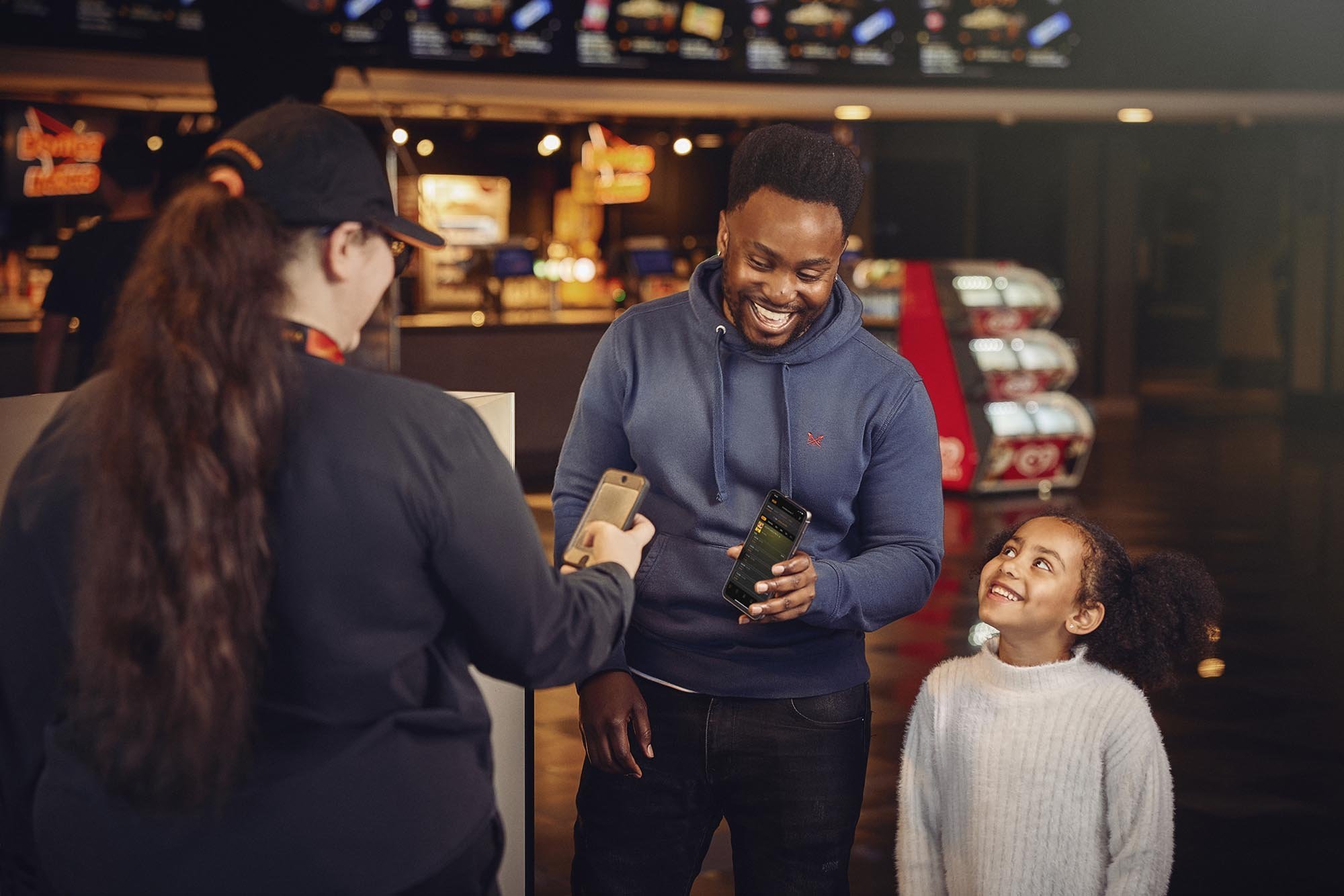 A dad and his daughter use their phone to show their cinema ticket to a Vue cinema staff member in this image by advertising photographer Jon Enoch