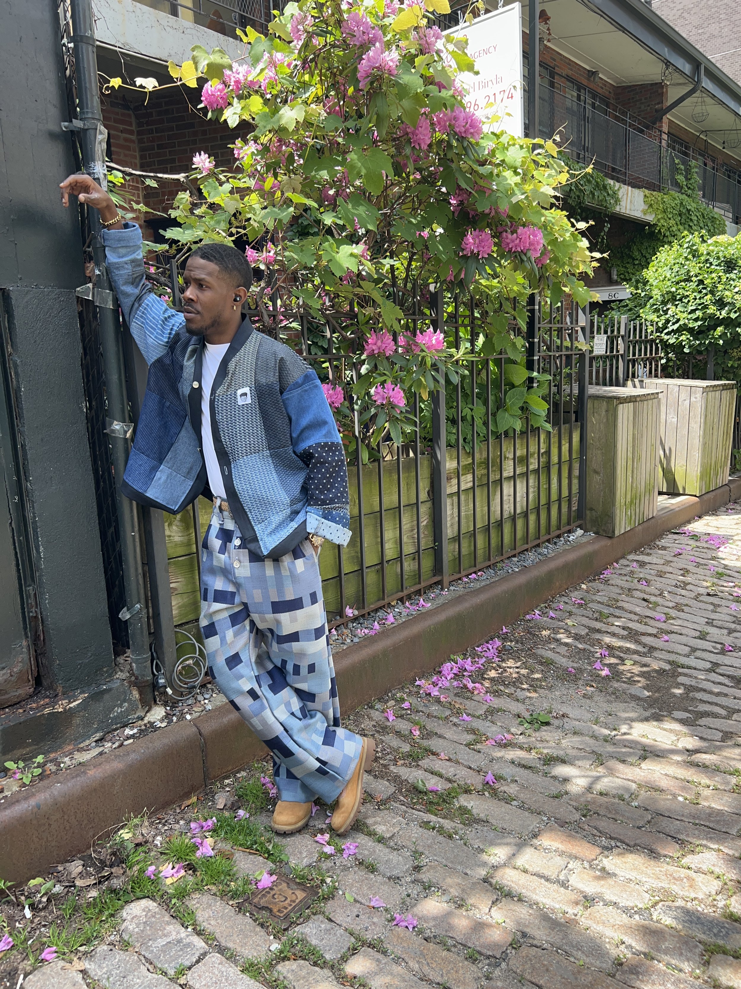 A man standing on a cobblestone sidewalk next to a black metal fence with pink flowering plants behind it. The man is wearing a multicolored jacket, plaid pants, yellow boots, and earbuds, and leans against a black wall.