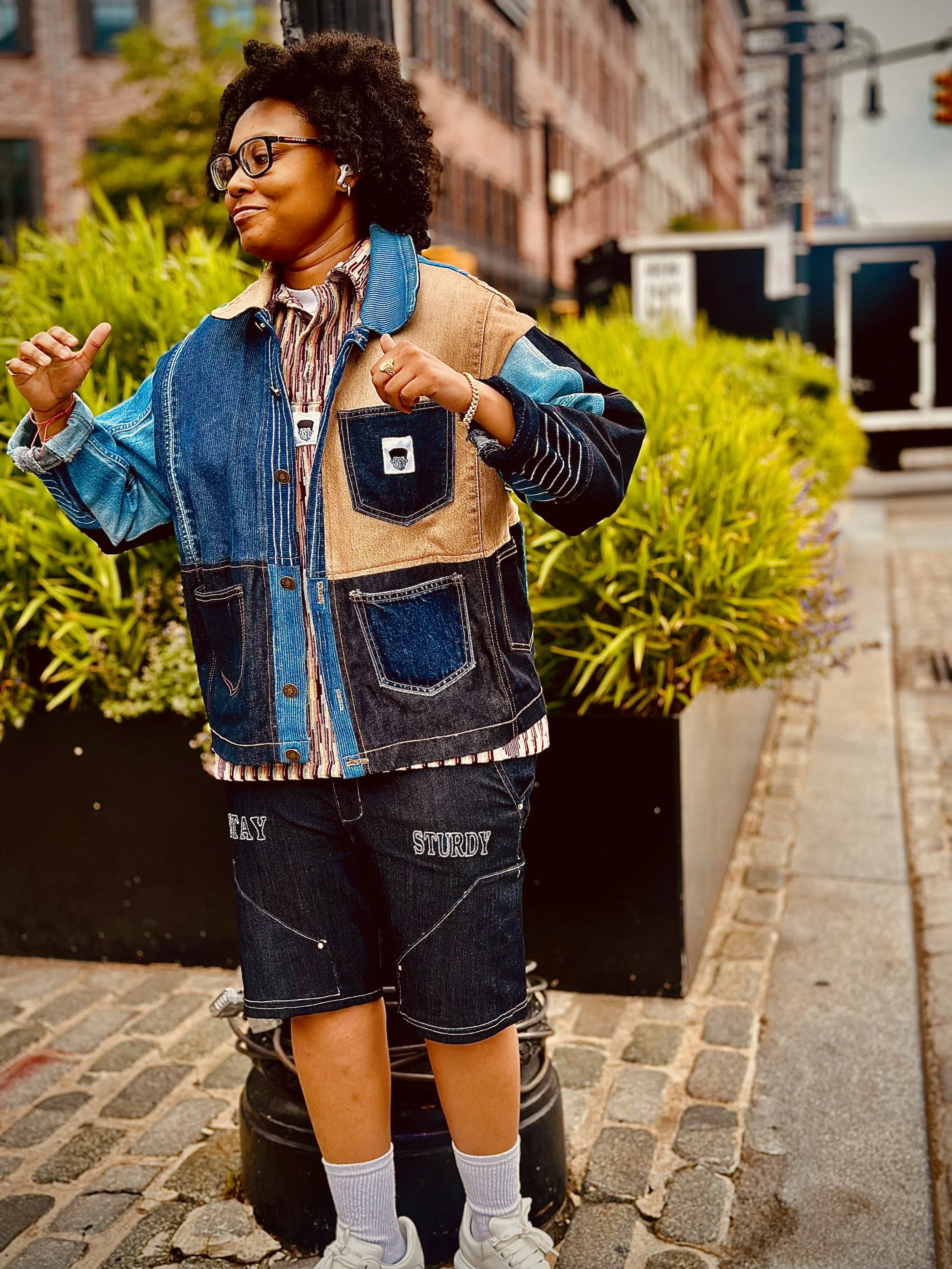 A person with curly hair wearing glasses, a multi-colored patchwork jacket, dark shorts with the words 'TAY' and 'STURDY' embroidered on them, white socks, and sneakers, standing on a cobblestone street with yellow plants and brick buildings in the background.