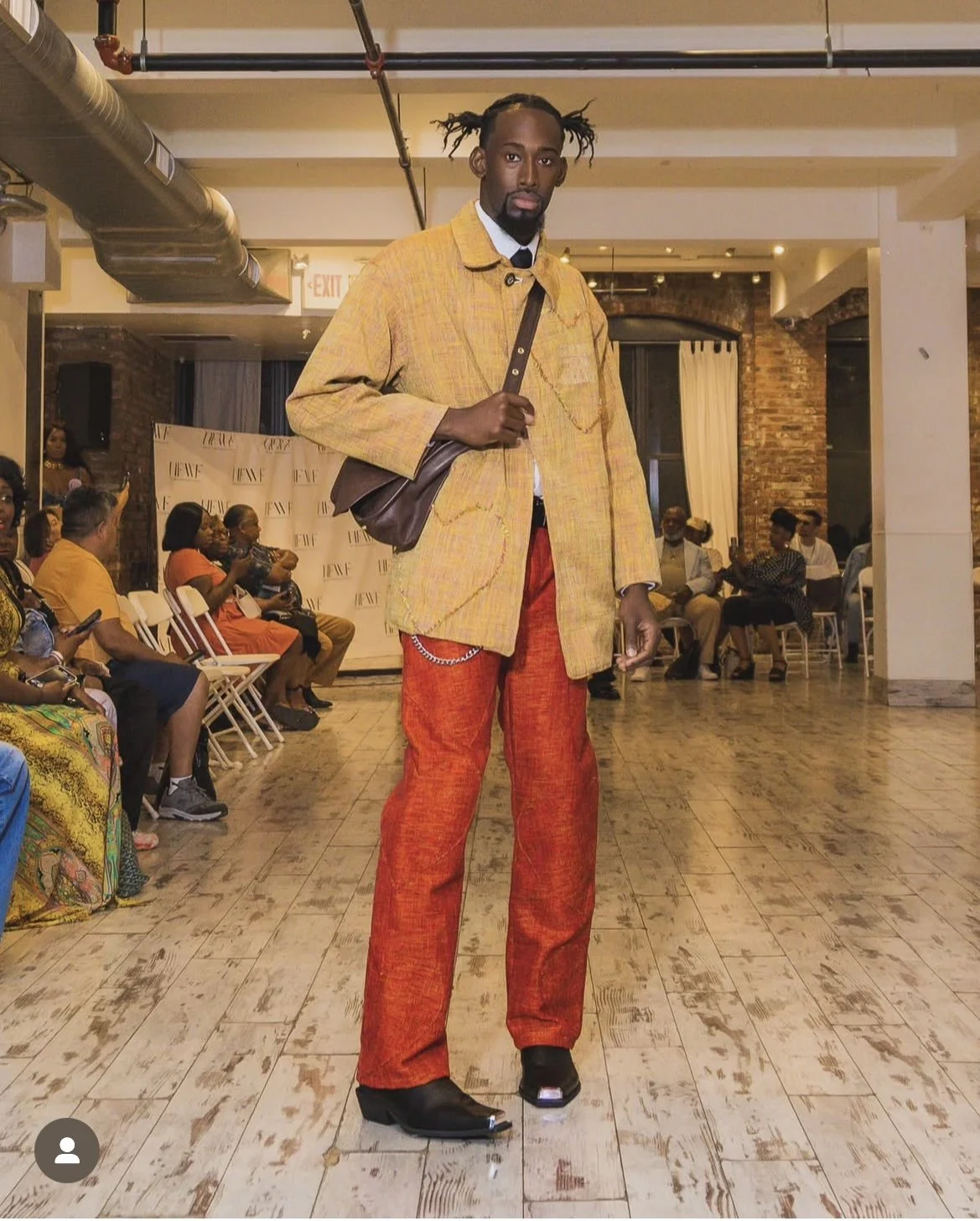 A man with styled hair and a beard stands in the center of a room with a wooden floor, wearing a yellow jacket, red pants, and black shoes, carrying a brown shoulder bag. People are seated on chairs along the walls, watching him, with some using their phones.