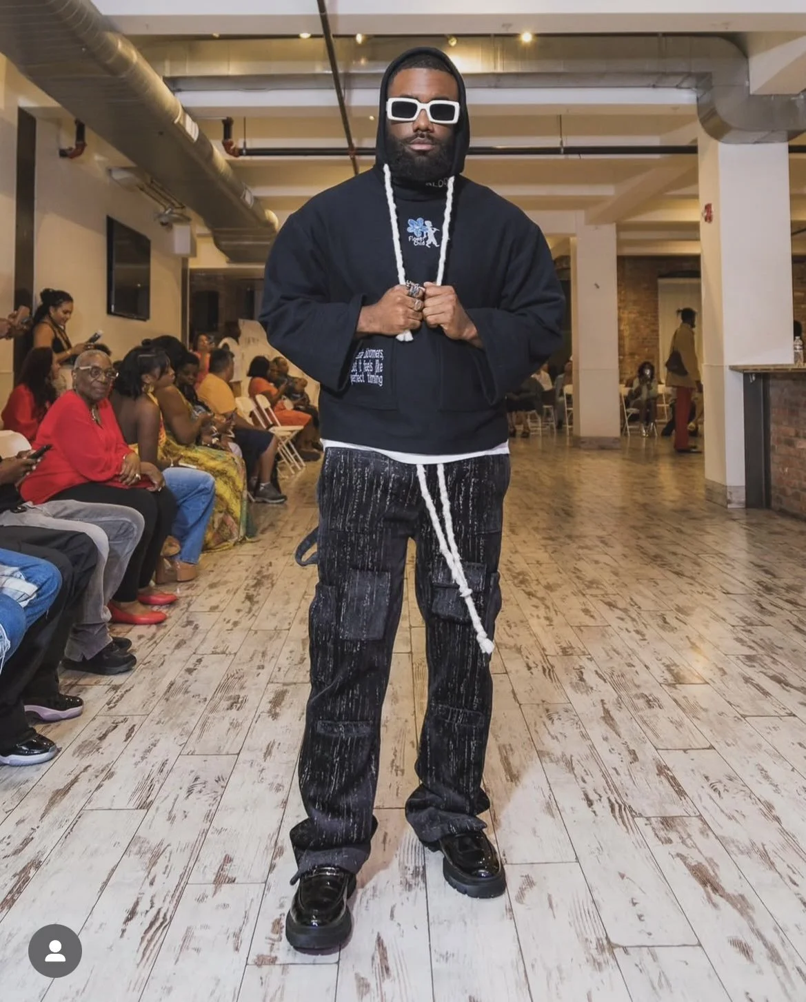 A man with a beard, wearing white sunglasses, a black hoodie and black and white paint-splattered pants, stands in the center of a room with wooden floors and exposed ductwork, facing the camera. He is holding a white rope with both hands. In the background, people are seated and standing, some looking at their phones, in a spacious, modern room with industrial decor.