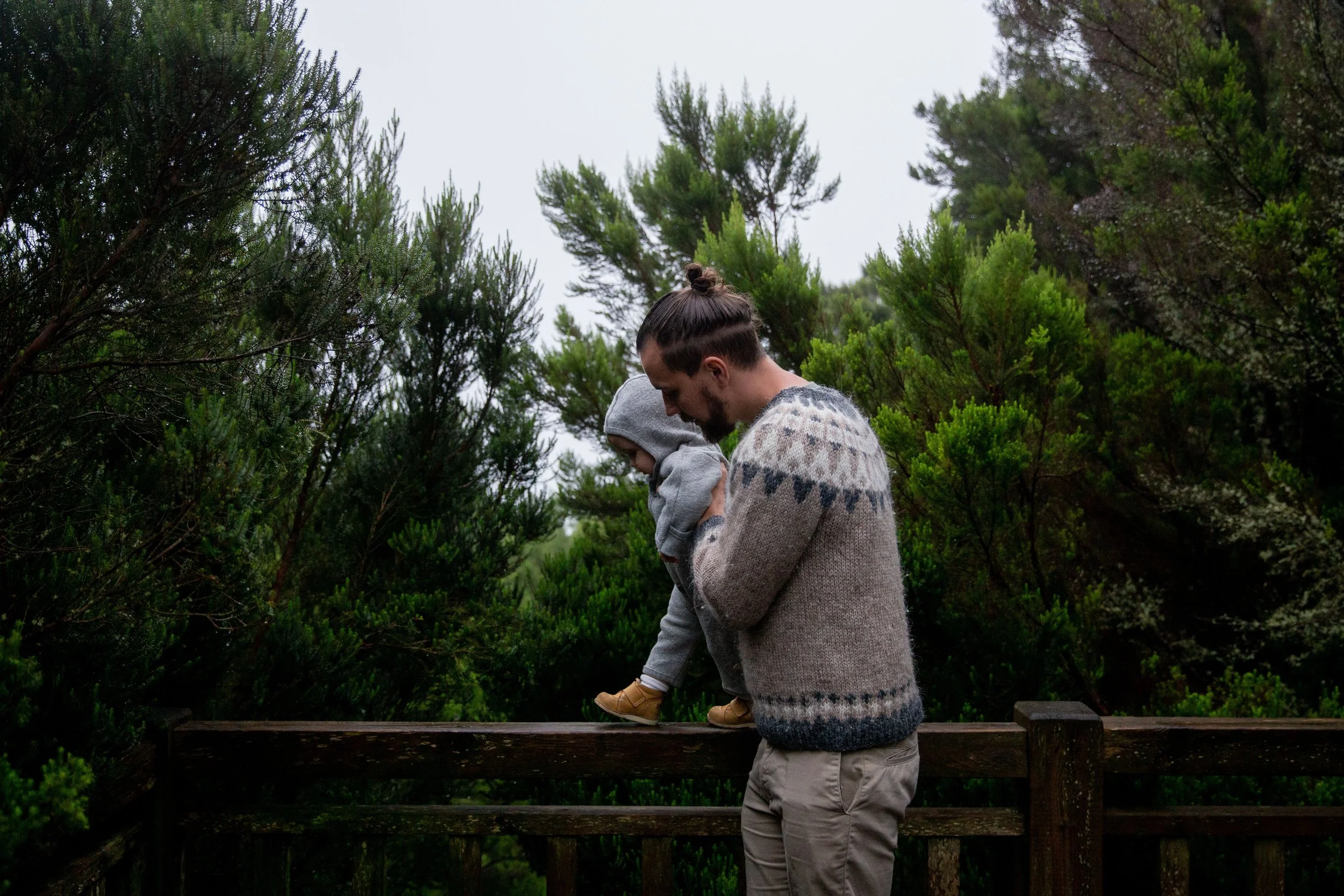 A man holding a young child, both wearing sweaters, standing on a wooden platform surrounded by lush green trees