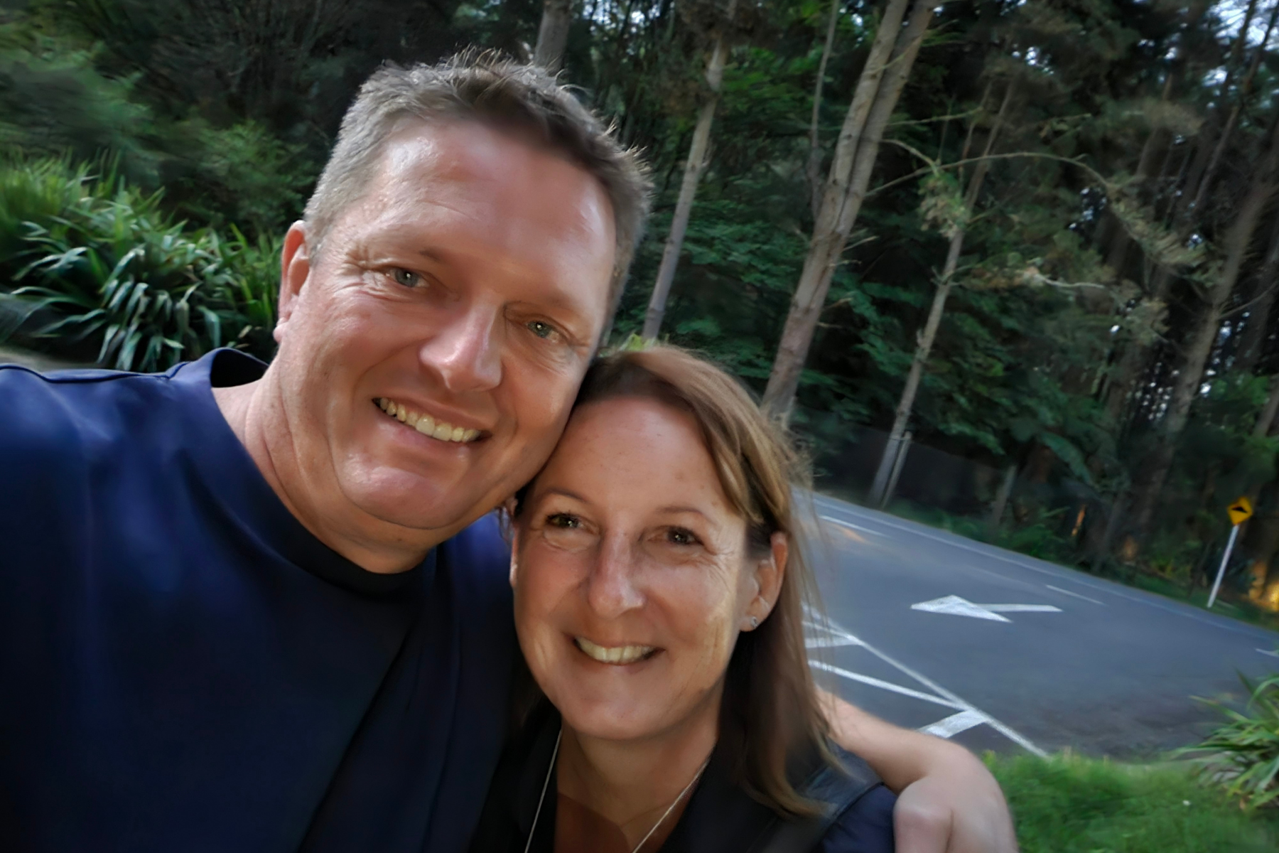 Daniel and his wife taking a selfie outdoors on a road surrounded by green trees.