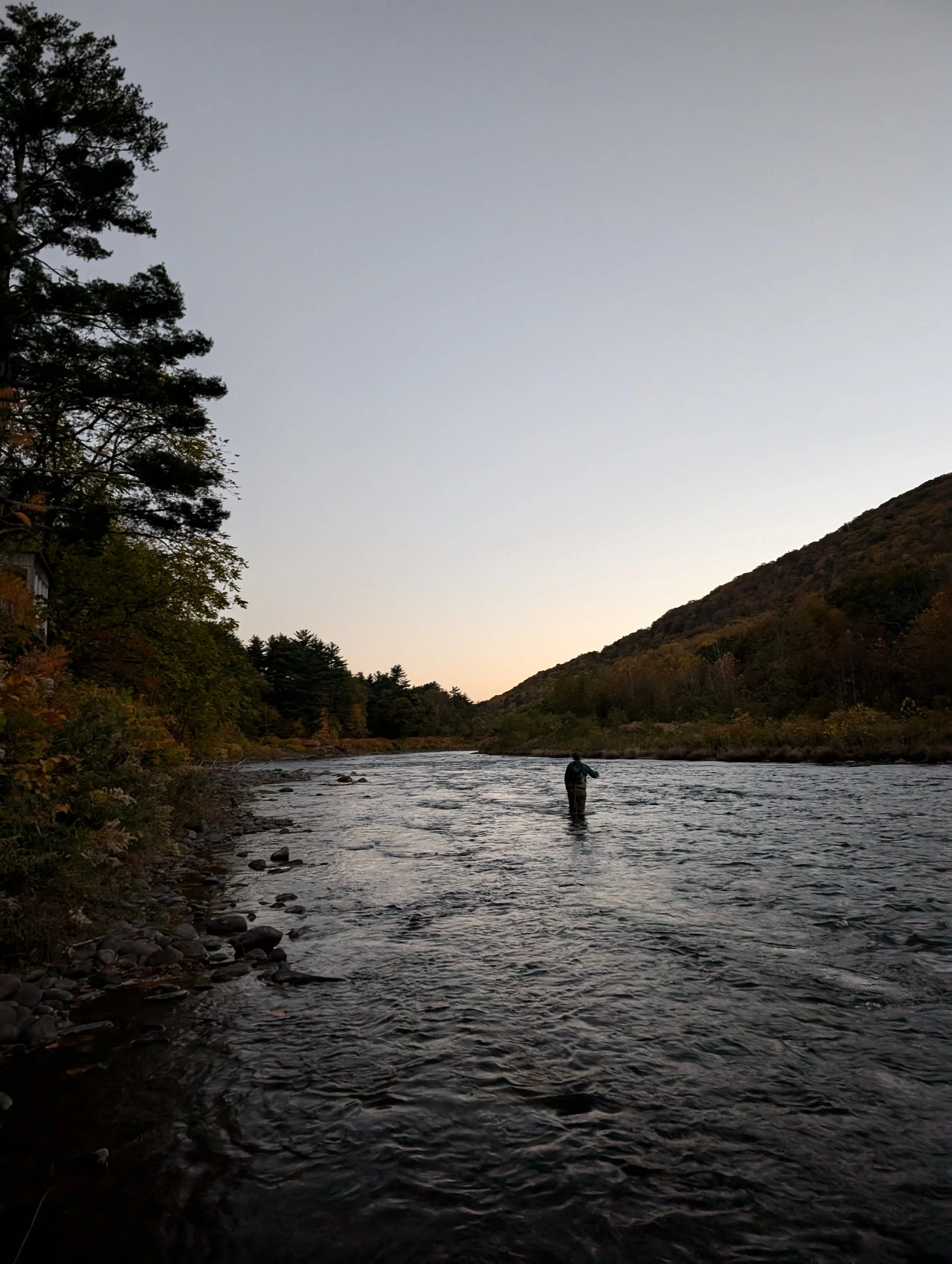 A person standing in a river at dusk, surrounded by forested hills and trees, with a clear sky above.