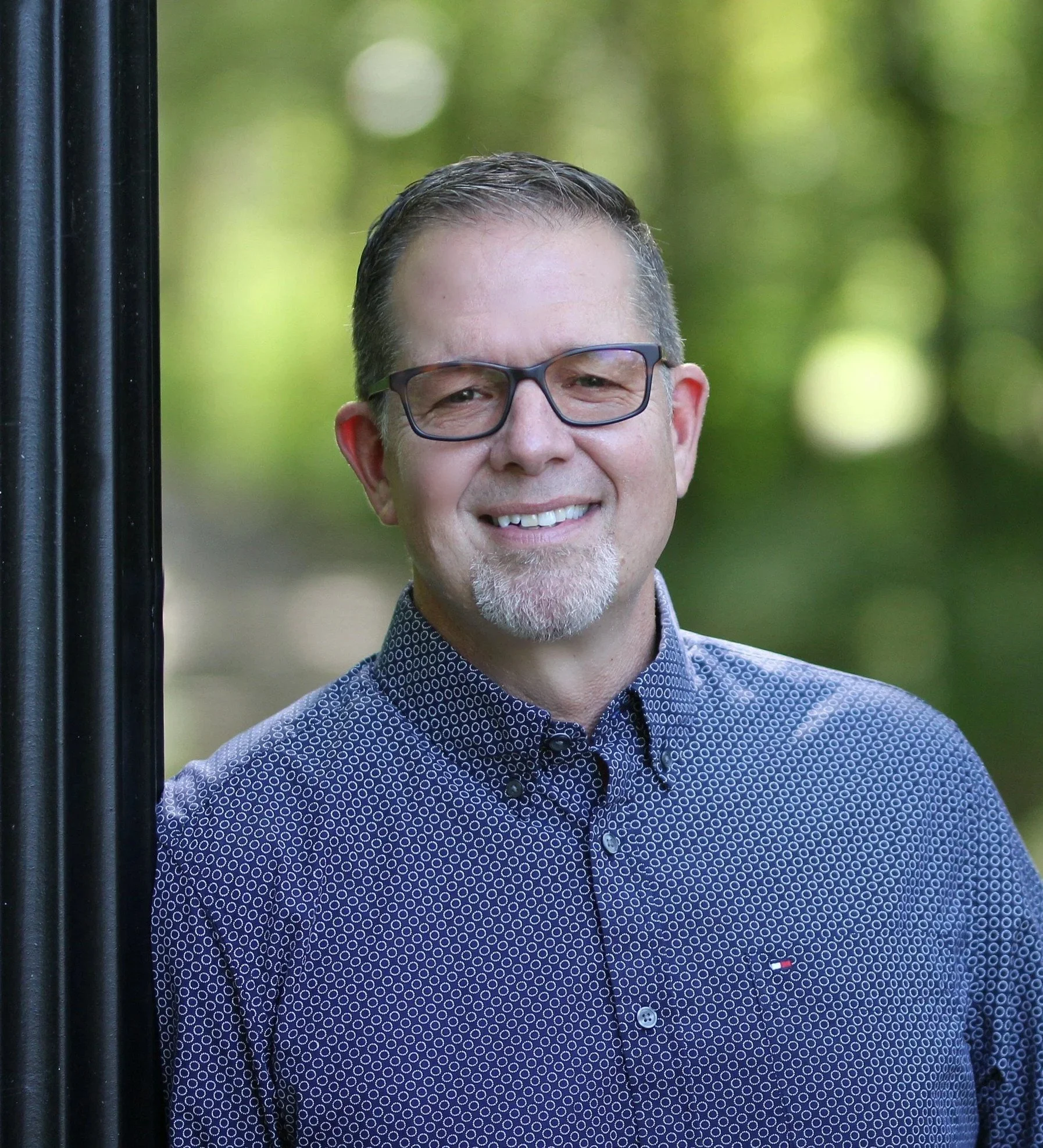 A smiling man with glasses, short gray hair, and a goatee, wearing a navy blue shirt with a small white pattern, standing outdoors with a blurred background of green trees.