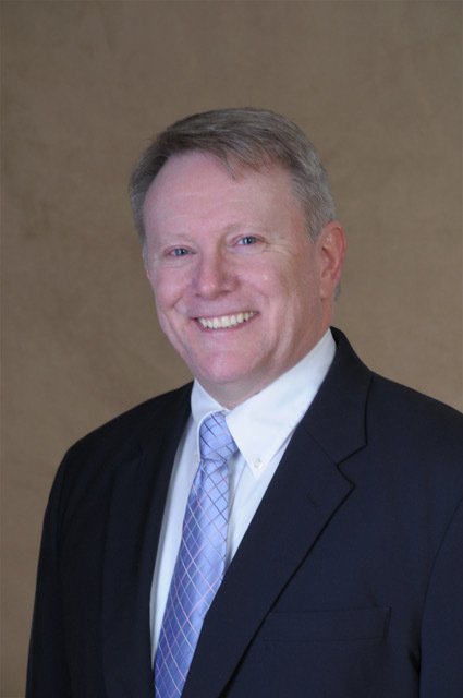 A smiling man in a dark suit, white shirt, and light purple tie, posing against a plain background.