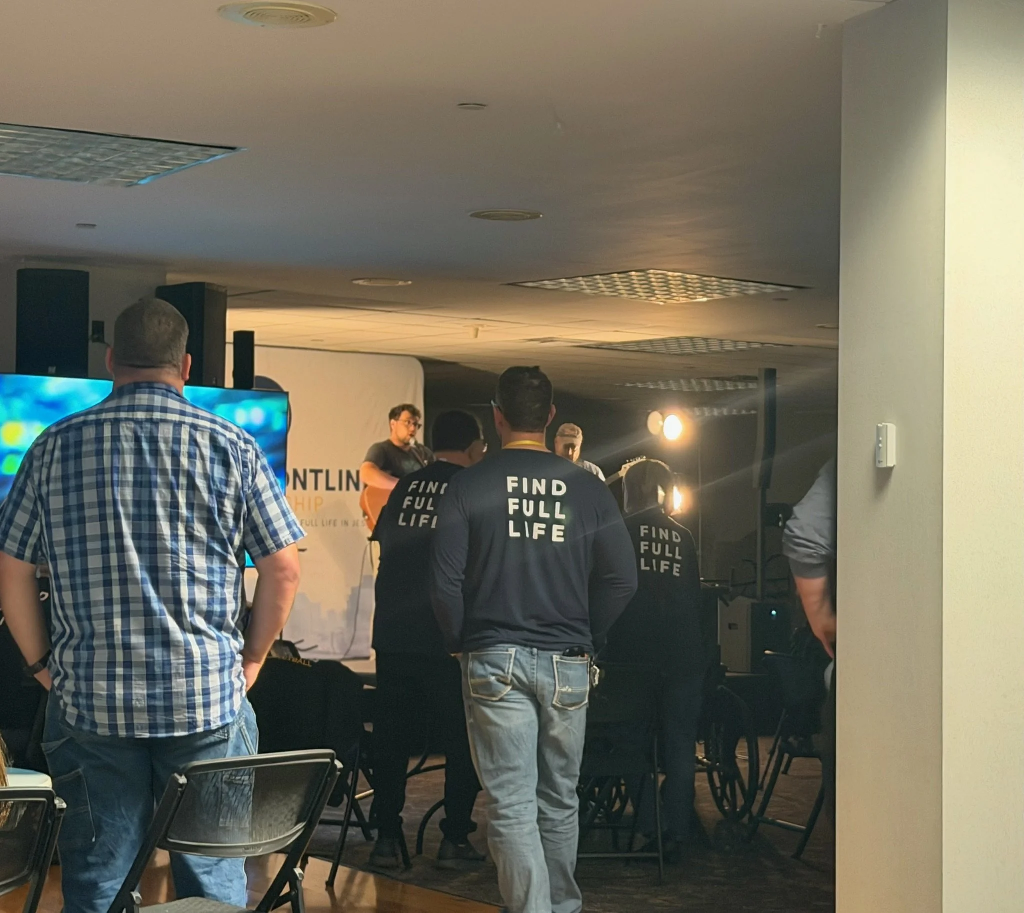 People standing and sitting in a room with a presentation screen, some wearing shirts that say "FIND FULL LIFE". The room has a ceiling with lights and ventilation panels, and there are chairs arranged in rows.