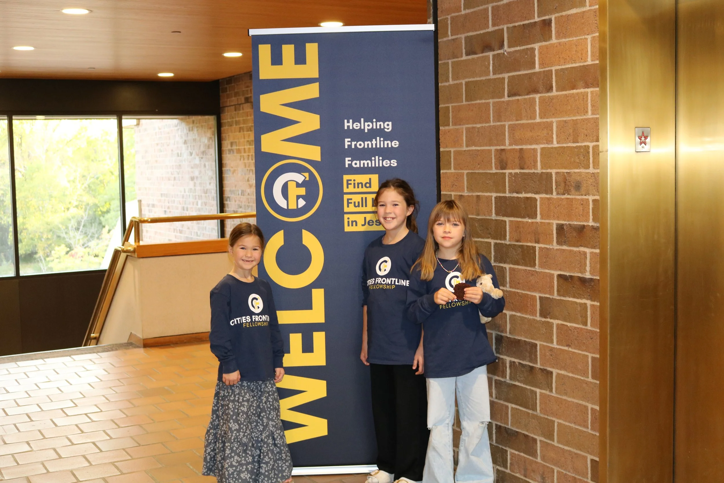 Four young girls standing in front of a blue banner that reads 'Help' and 'Helping Frontline Families' with a logo. The girls are smiling and wearing navy blue sweatshirts with the same logo, indoors near a brick wall and glass window.