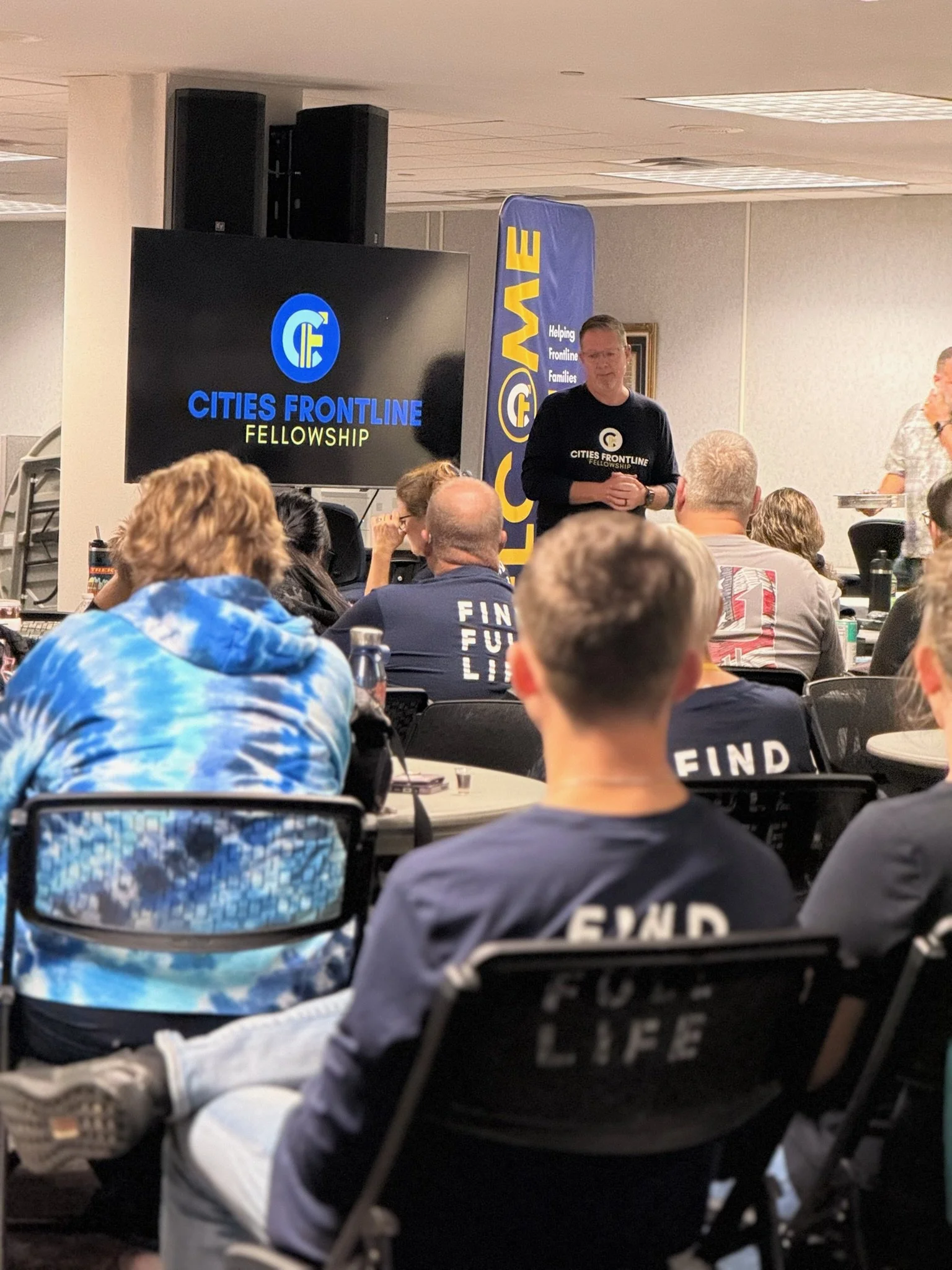 A man standing in front of a screen and a banner at a workshop or seminar, with an audience seated in a room.