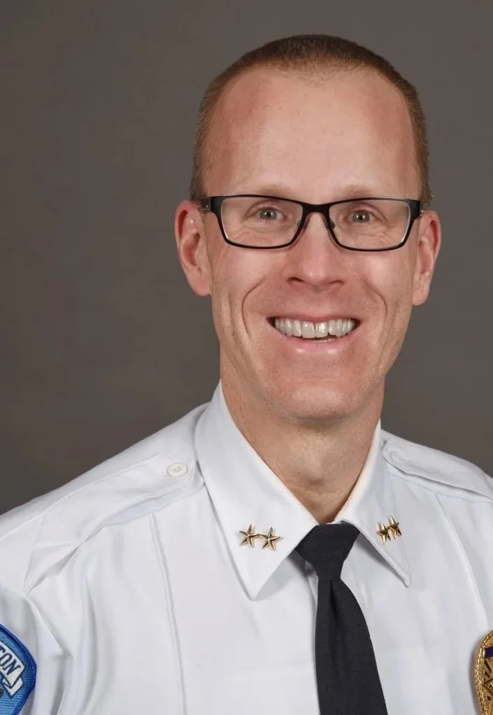 A smiling man in a white police uniform with gold star insignias on his collar, black glasses, and short red hair, posing against a gray background.