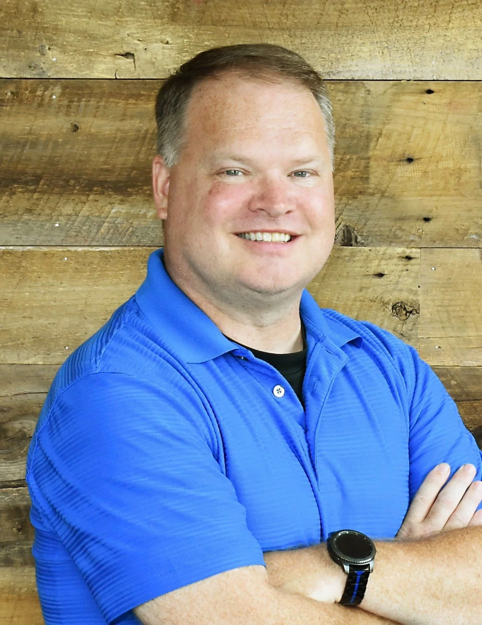 Portrait of a man in a blue shirt, smiling, with a wooden background.