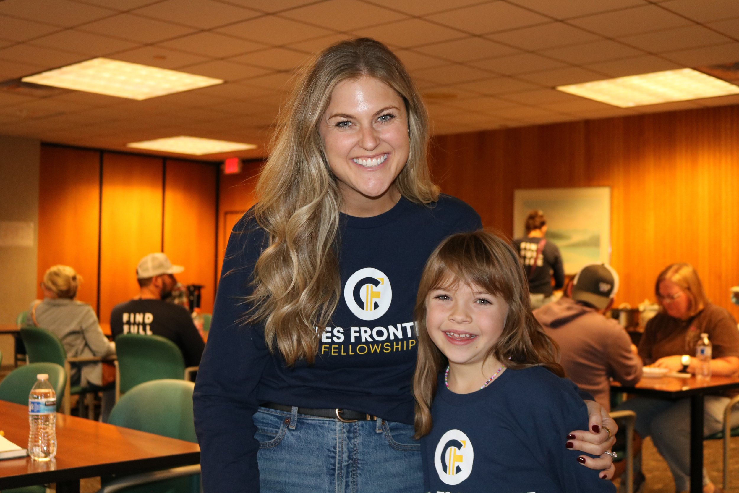 Two smiling women, one older with long wavy blonde hair and one younger with shoulder-length light brown hair, wearing matching navy blue shirts with a circular logo and the words "FACES FRONT FELLOWSHIP," are standing together in a conference room with people seated at tables in the background.