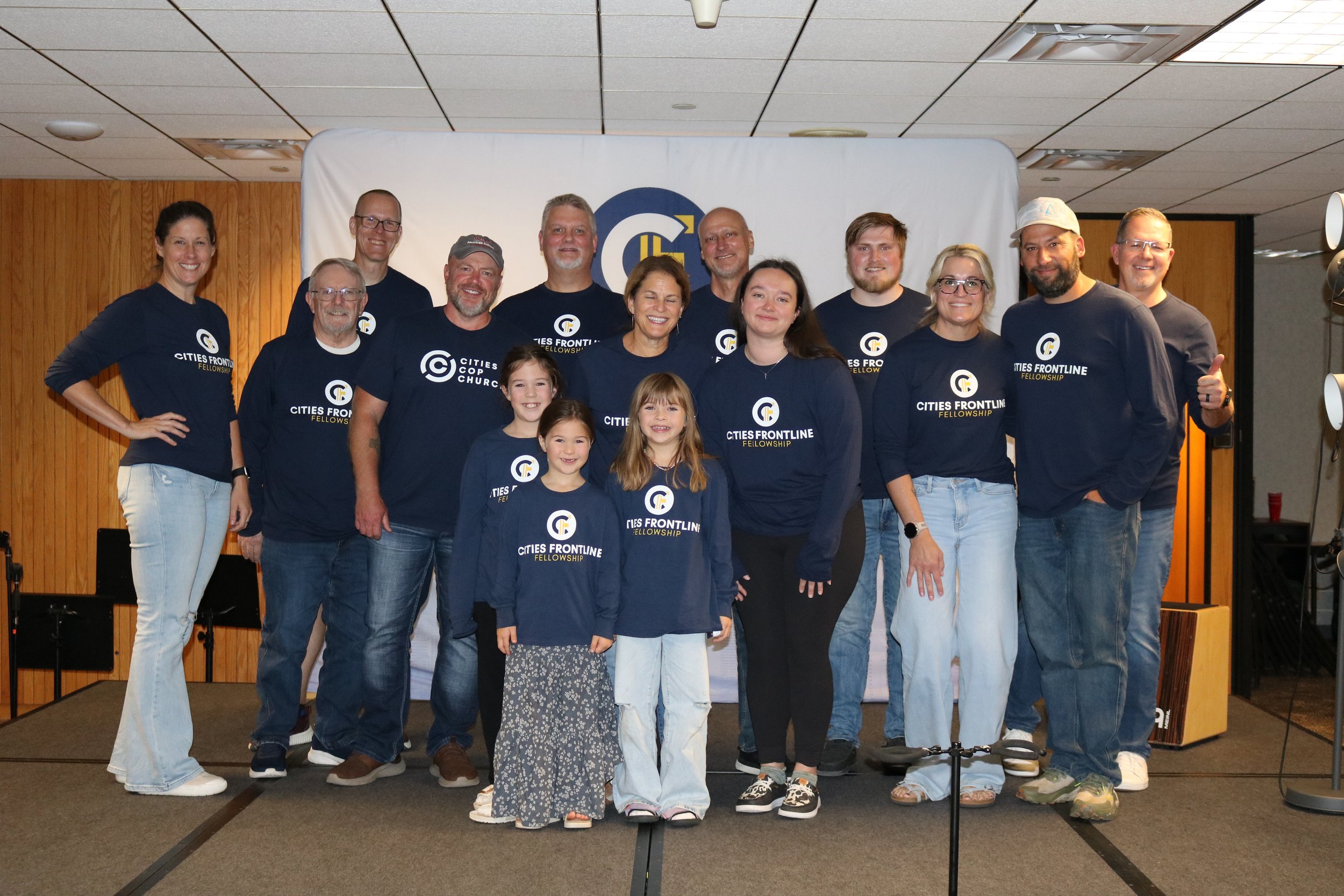A group of 14 people, including children, posing together on a stage with a white backdrop featuring a logo and the words "Cities Frontline Fellowship." They are wearing matching navy blue shirts with the same logo and text, smiling for the photo.