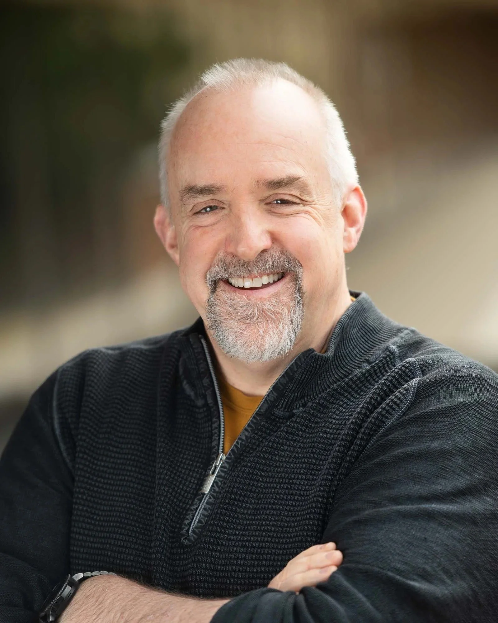 Smiling older man with gray hair and beard, wearing a black zip-up jacket, sitting outdoors with arms crossed.