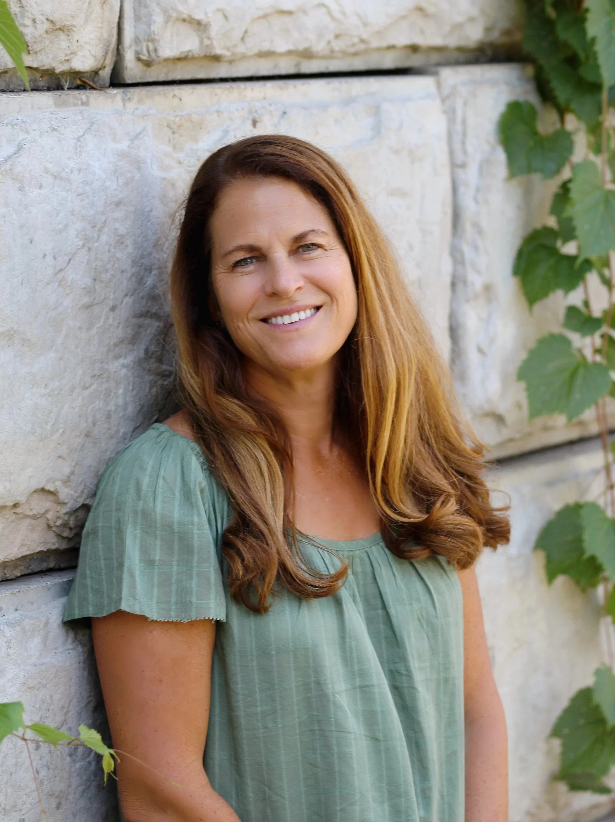 A woman with long, wavy brown hair and a bright smile standing against a stone wall with green foliage on the right.