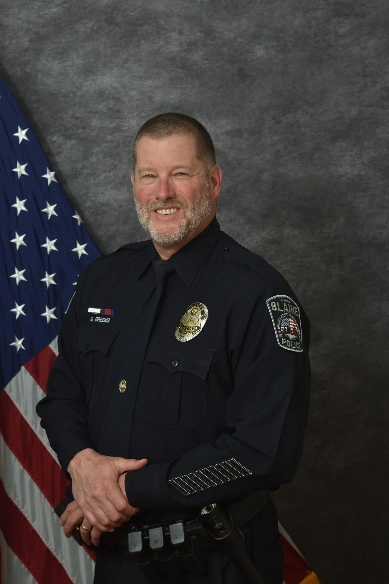 Portrait of a male police officer with a beard, wearing a dark police uniform, standing in front of an American flag and gray background, smiling.